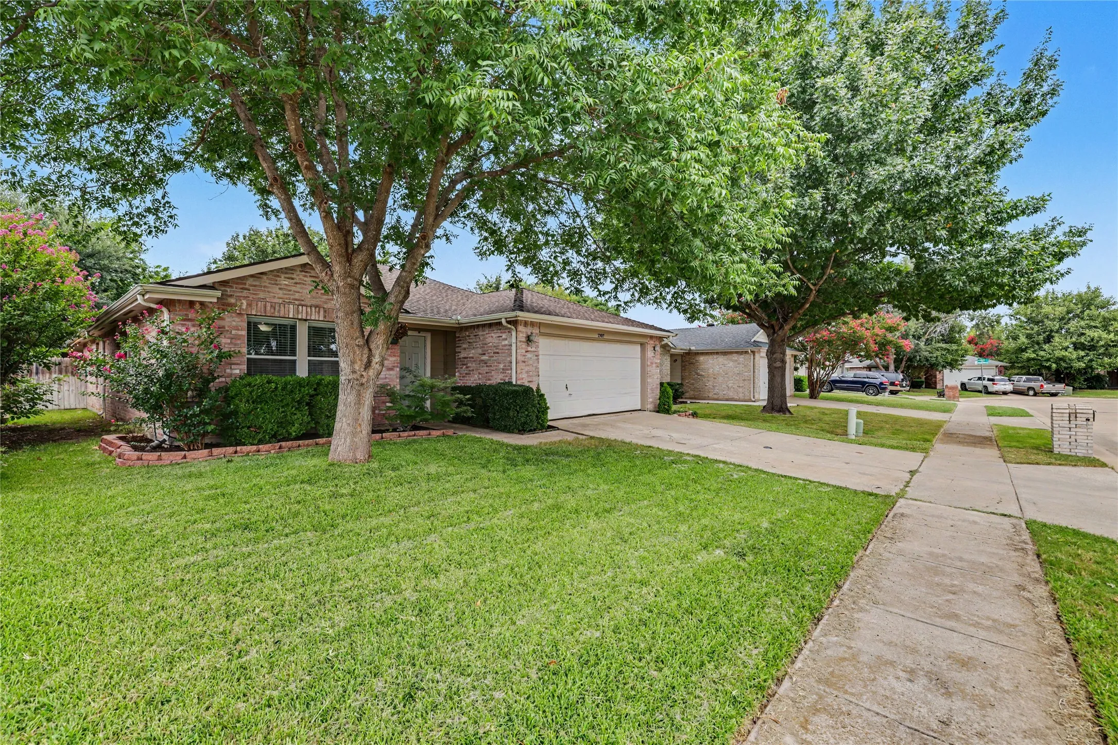Brick exterior home with an attached two-car garage, mature front yard trees, a concrete driveway, and a sidewalk