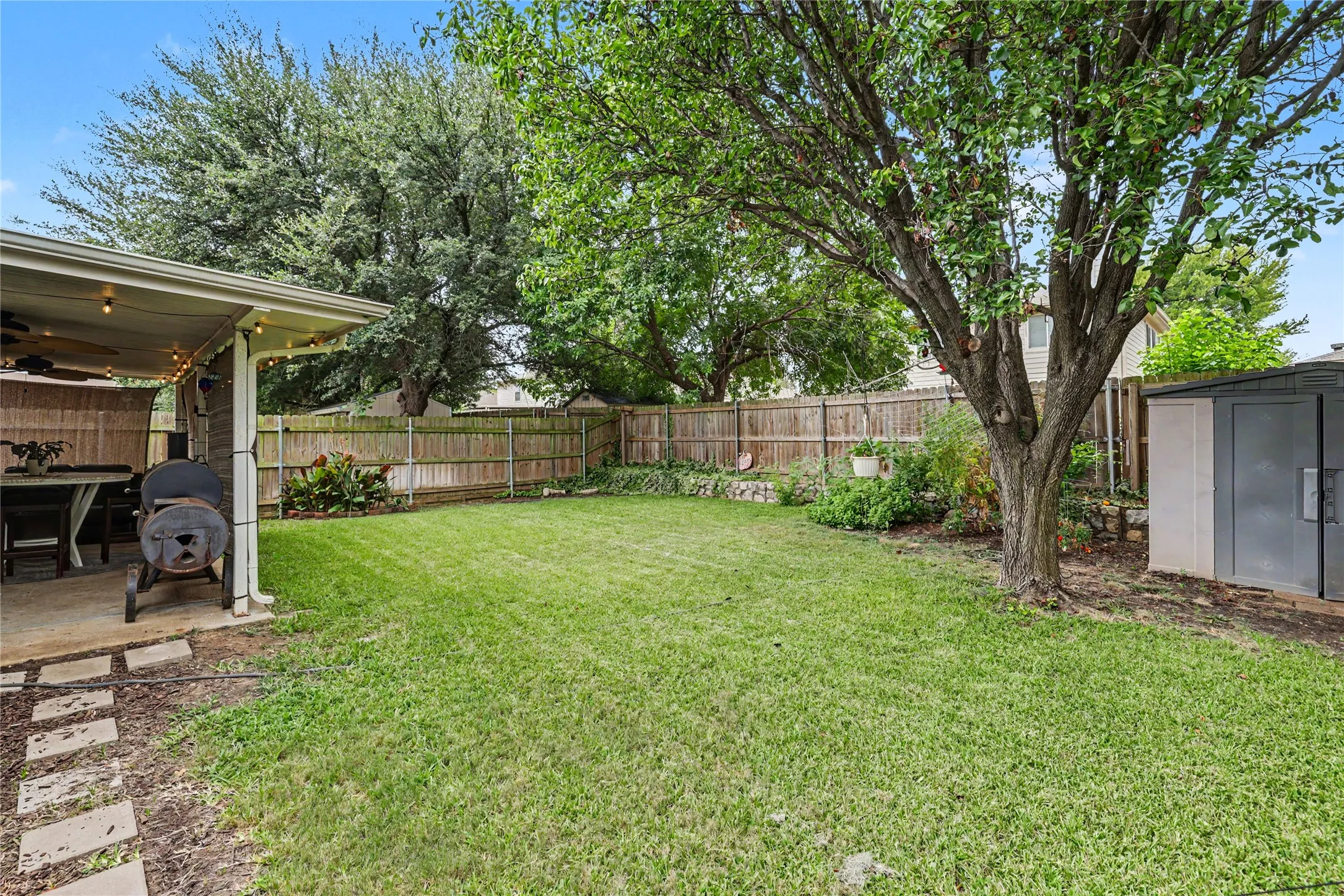 Expansive backyard featuring a covered patio with string lighting, a large grassy area, mature trees, a privacy fence, and a storage shed