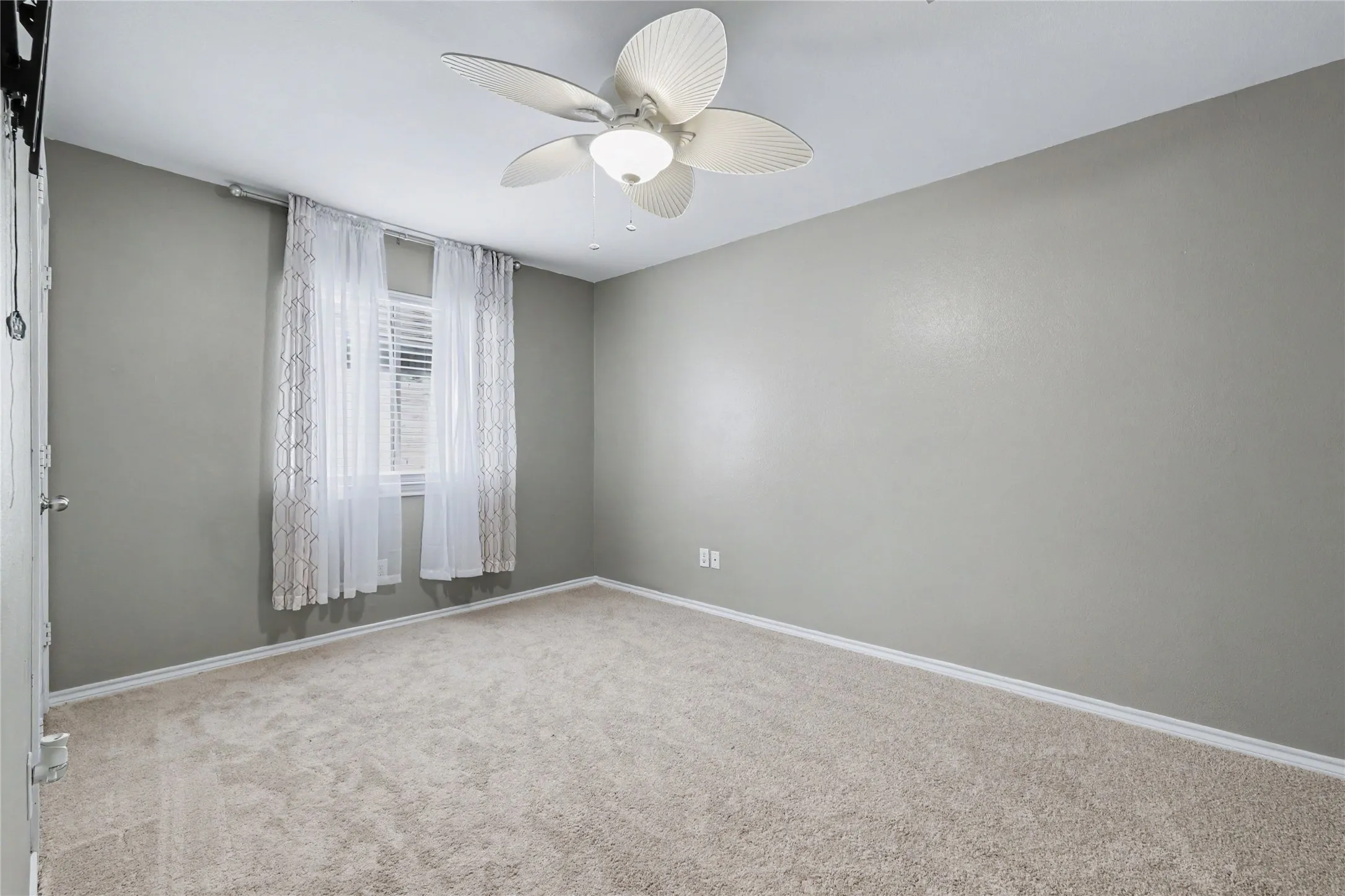 Carpeted interior space featuring a window with horizontal blinds and decorative sheer curtains, a ceiling fan with integrated lighting, and neutral wall tones