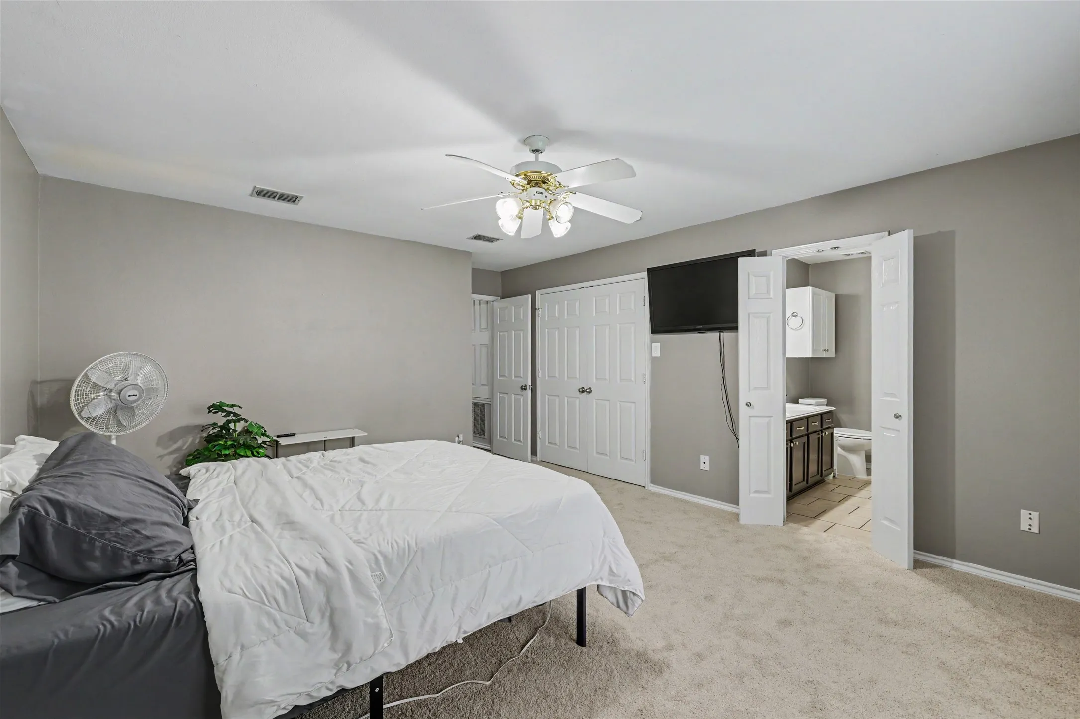 Carpeted room featuring a ceiling fan with integrated lighting, two bi-fold closet doors, and an en-suite bathroom entrance with a vanity and toilet
