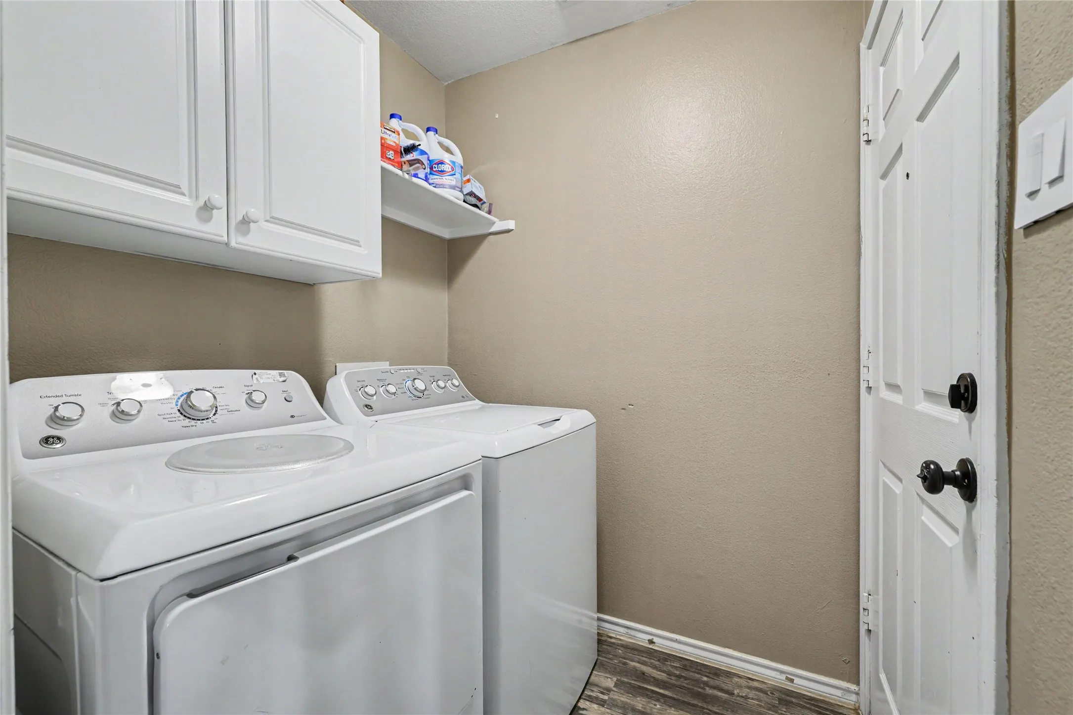 Dedicated laundry area featuring white upper cabinetry, a coordinating white wall shelf, and wood-finish flooring