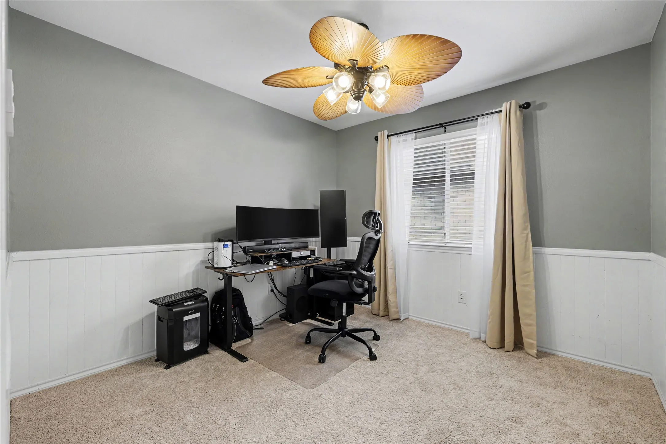 Interior room featuring light gray walls with white wainscoting, a window with blinds, and a ceiling fan with light fixture