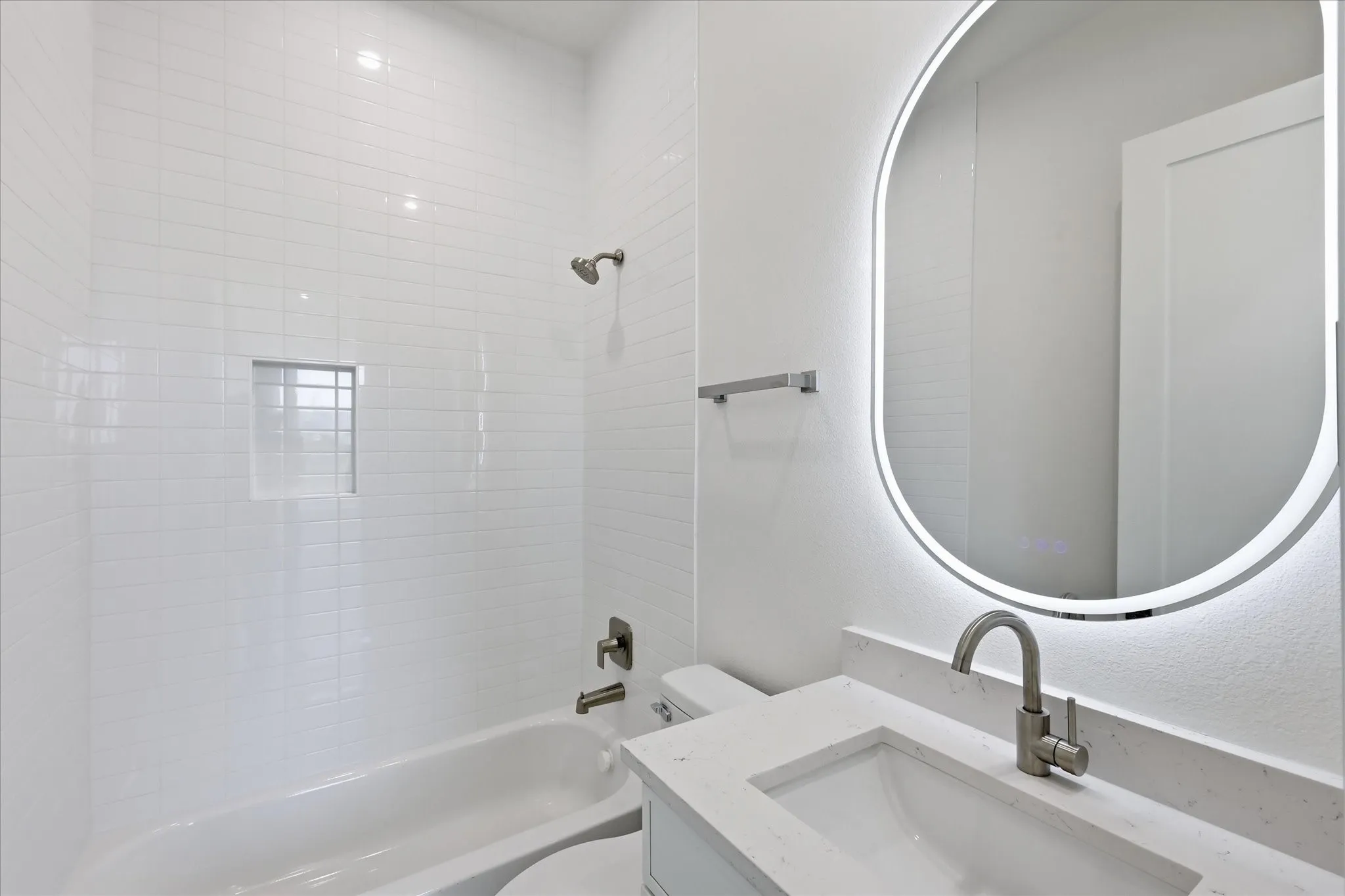 Bathroom featuring a white subway tile shower, built-in shower niche, brushed nickel fixtures, white vanity with an integrated sink, and an illuminated oval mirror