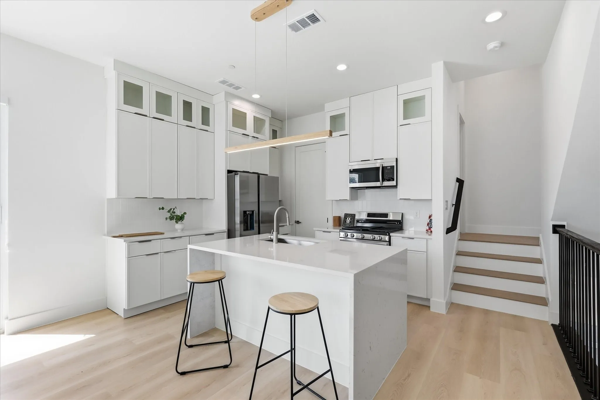Open-concept kitchen featuring an island with a waterfall countertop, integrated stainless steel appliances, abundant white cabinetry with some glass-front uppers, and wood-finish flooring