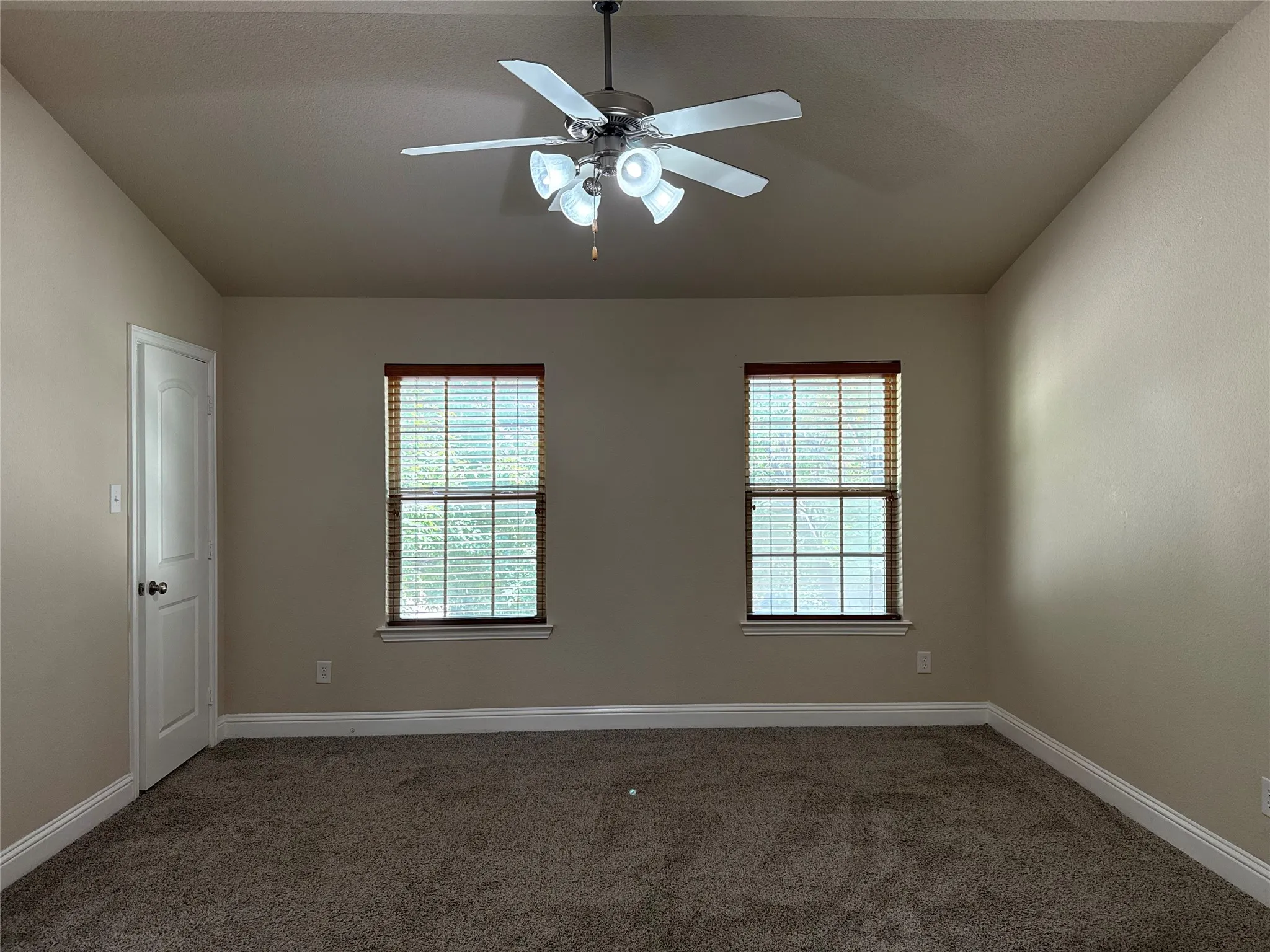 Neutral-toned room featuring a ceiling fan with integrated lighting, two double-hung windows with blinds, plush carpet flooring, and white baseboards