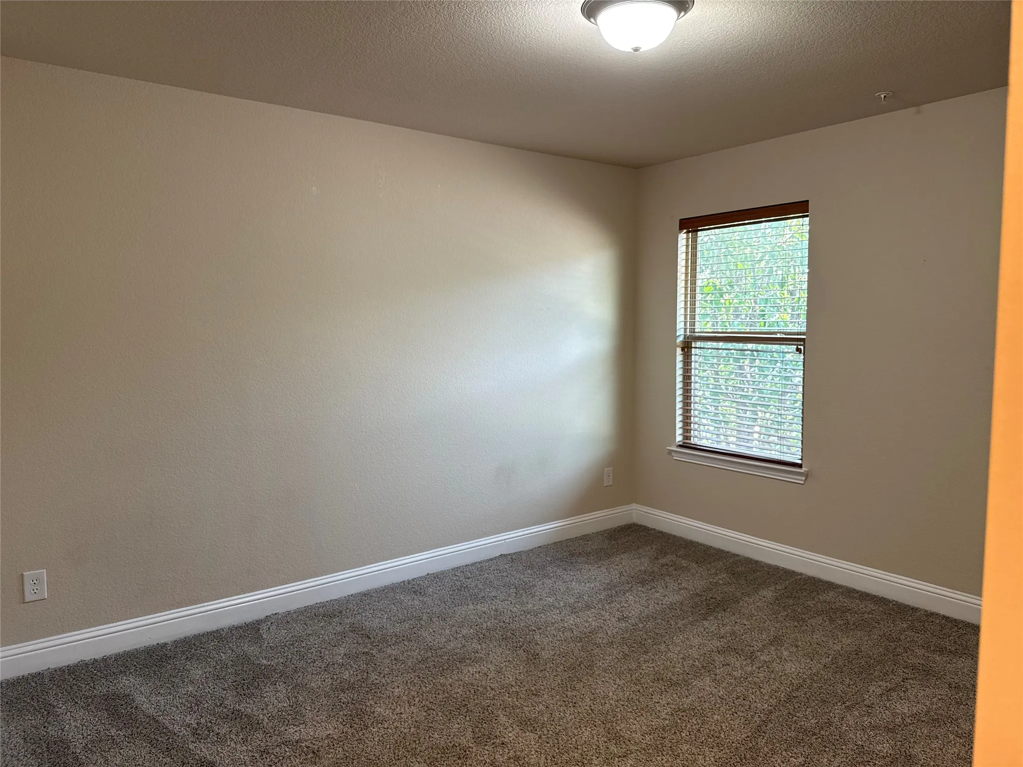 Neutral-toned room featuring a single window with blinds, overhead flush mount light fixture, textured walls, and carpet flooring