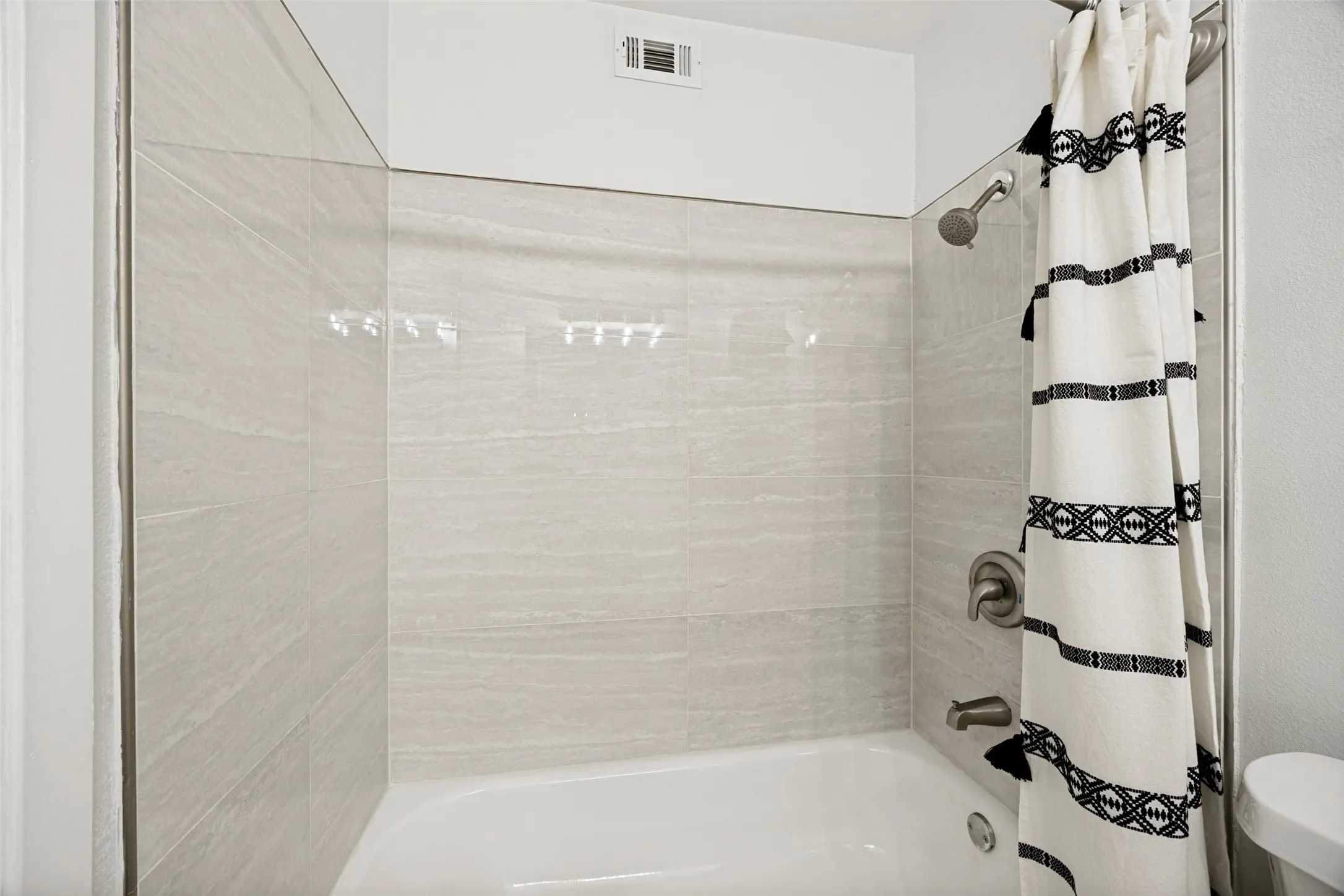 Bathtub with neutral-toned tiled surround, brushed nickel fixtures, and white ceiling with vent