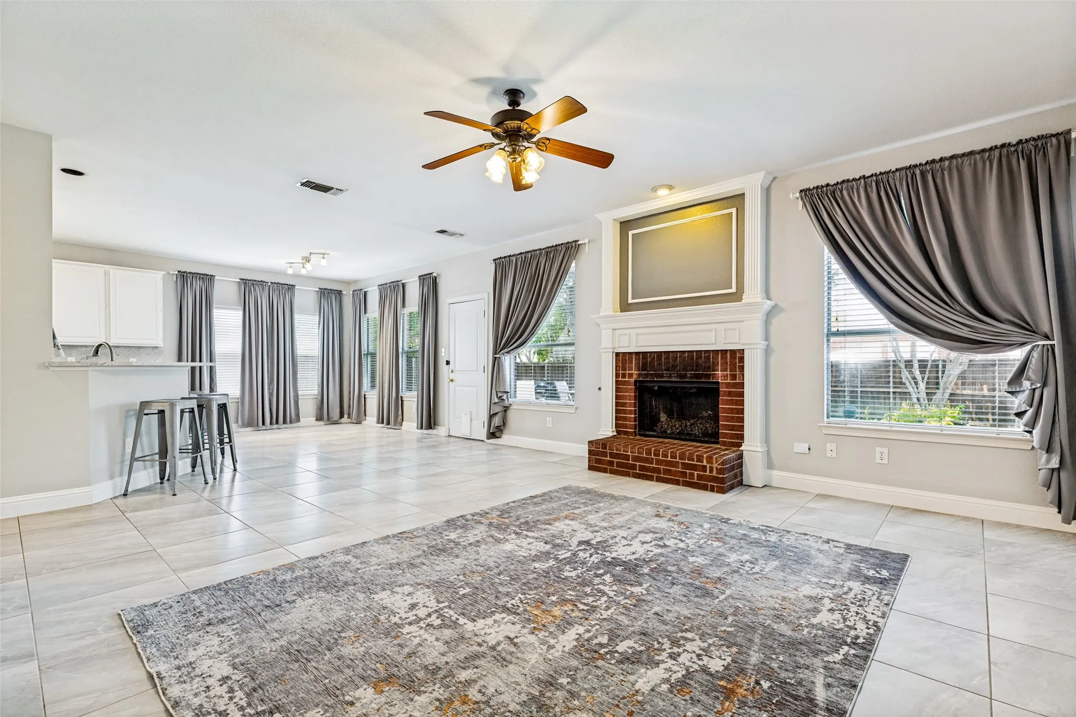 Spacious living area featuring a brick fireplace with a white mantel, ceiling fan, and light-toned tile flooring