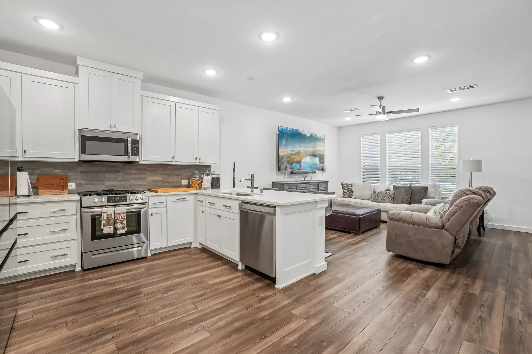 Open-concept living space featuring a kitchen with white cabinetry and stainless steel appliances, a center island with a sink, wood-finish flooring, and a living area with multiple windows