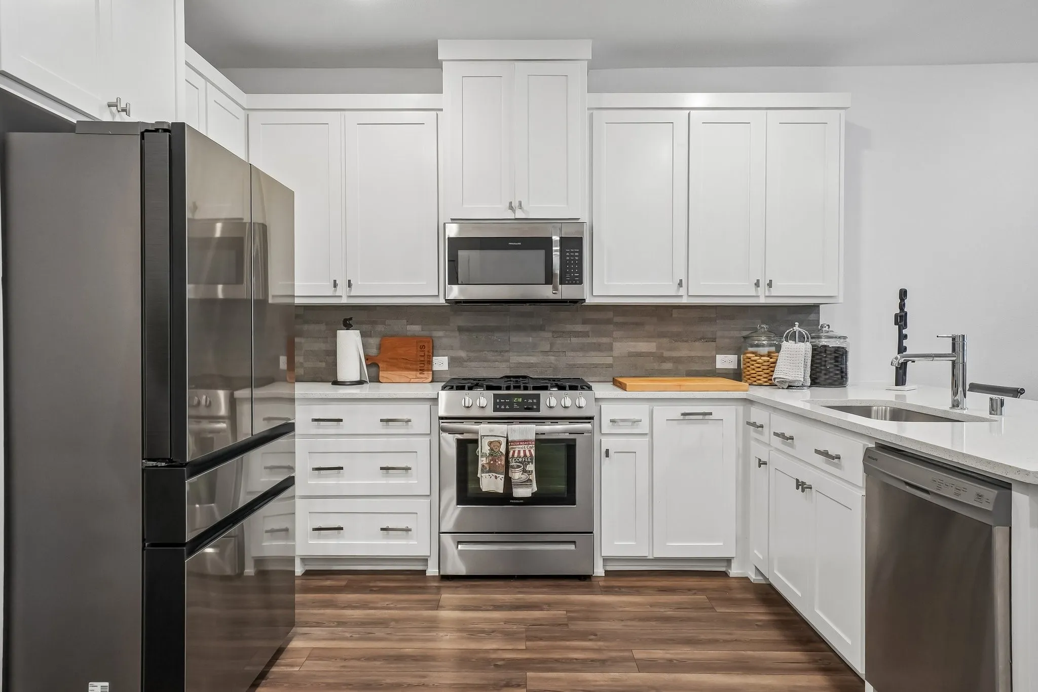 Modern kitchen featuring white shaker cabinetry, stainless steel appliances, a grey mosaic tile backsplash, white countertops, and wood-finish flooring