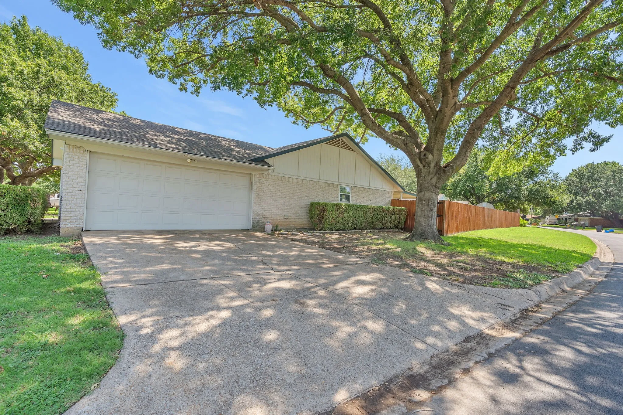 Exterior featuring a light brick façade, two-car garage, concrete driveway, and mature trees providing shade