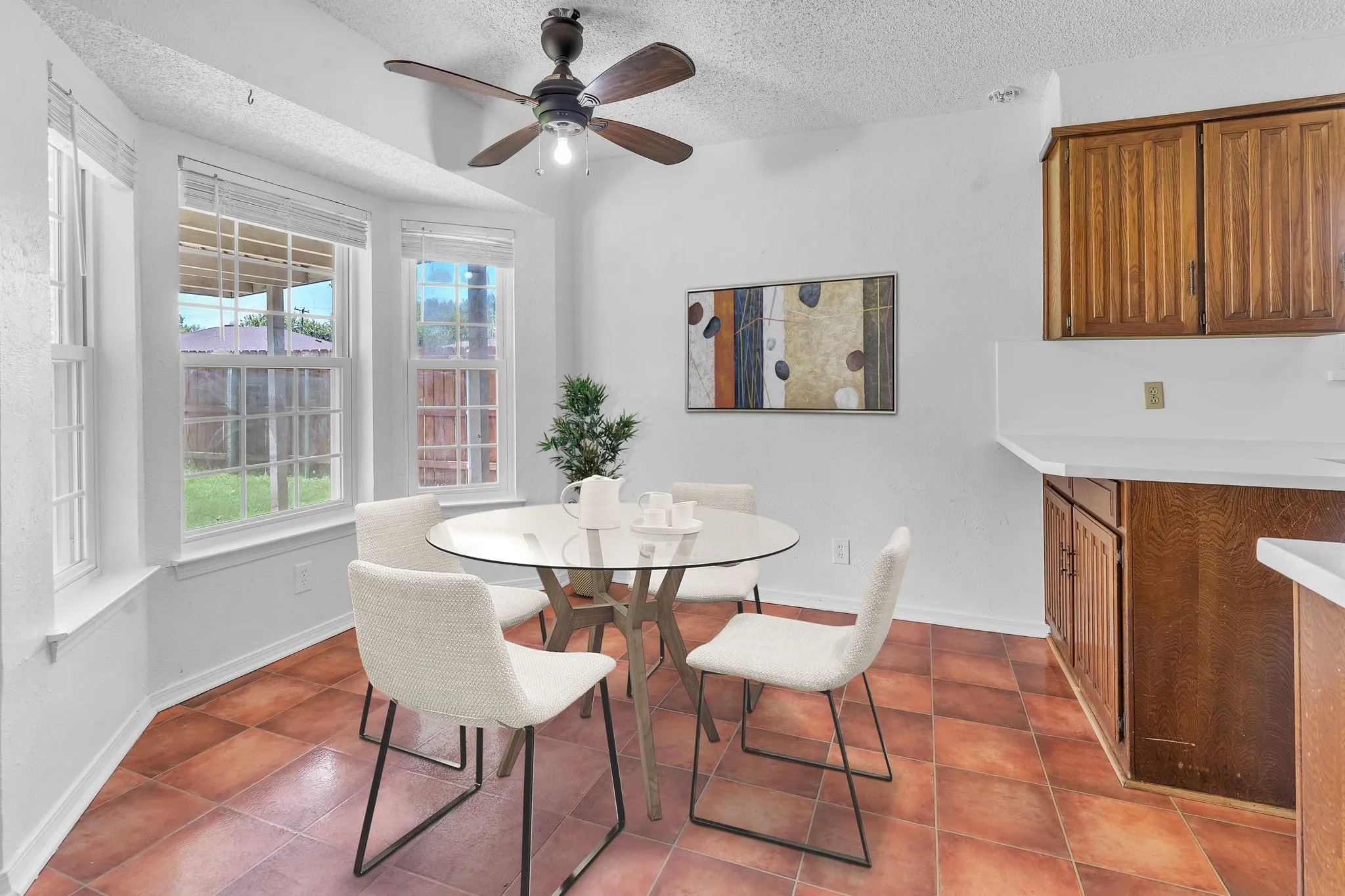 Bright dining area featuring a bay window, terracotta-style floor tiling, wood-finish cabinetry, white countertops, and a ceiling fan with integrated lighting