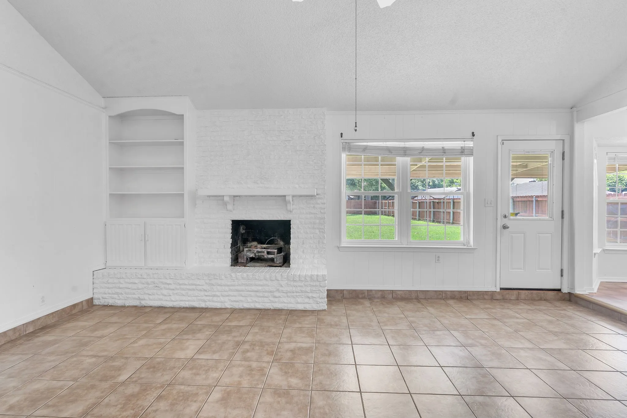 Spacious living area featuring tile flooring, a painted brick fireplace, built-in shelving with lower cabinetry, and ample natural light from multiple windows