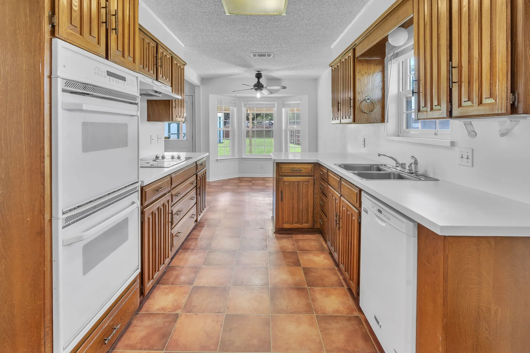 Kitchen featuring wood cabinetry, solid surface countertops, white appliances, a double wall oven, and a double basin sink