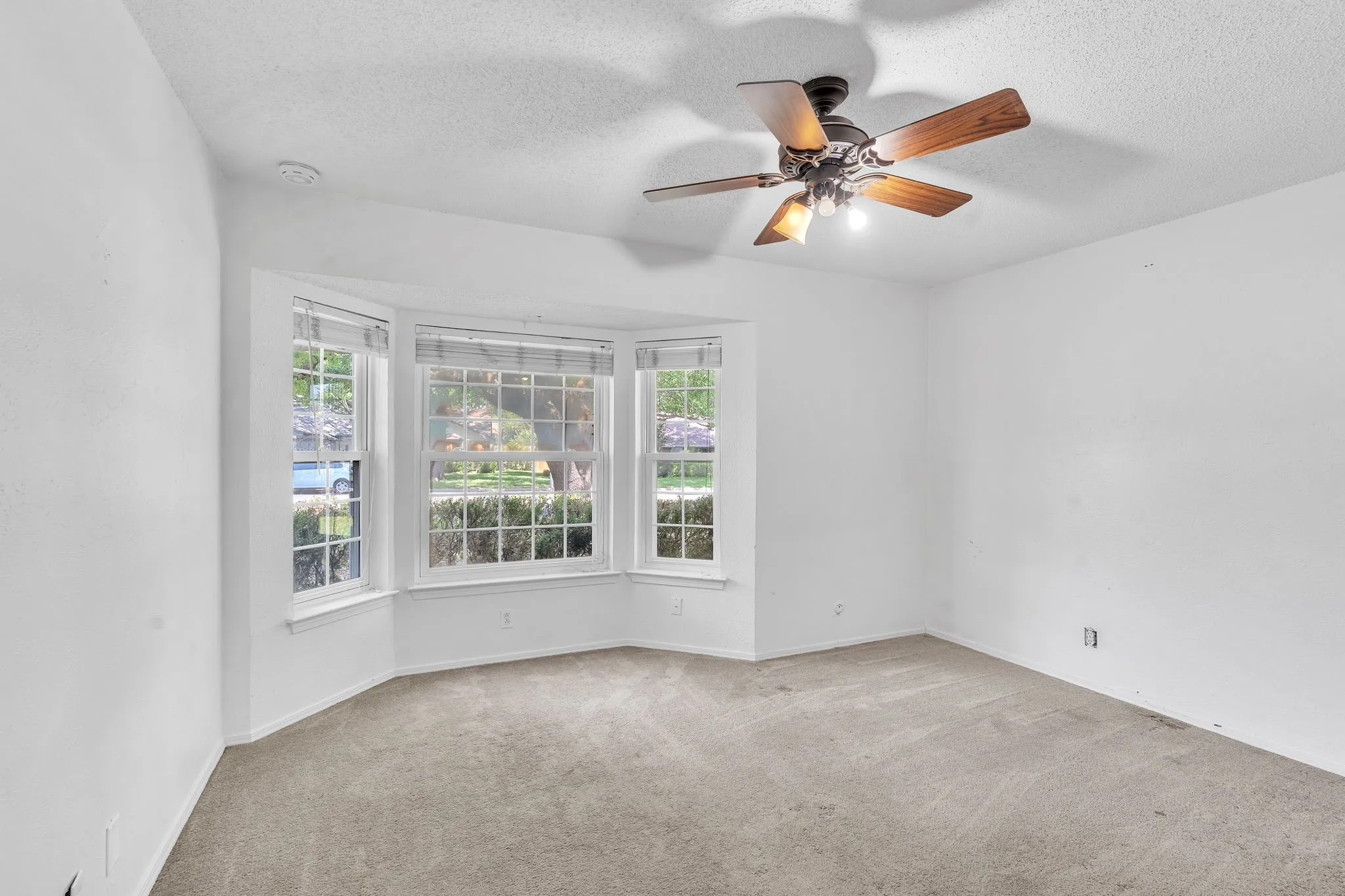 Spacious room featuring a bay window with multiple panes, ceiling fan with light fixture, textured ceilings, and neutral carpet flooring