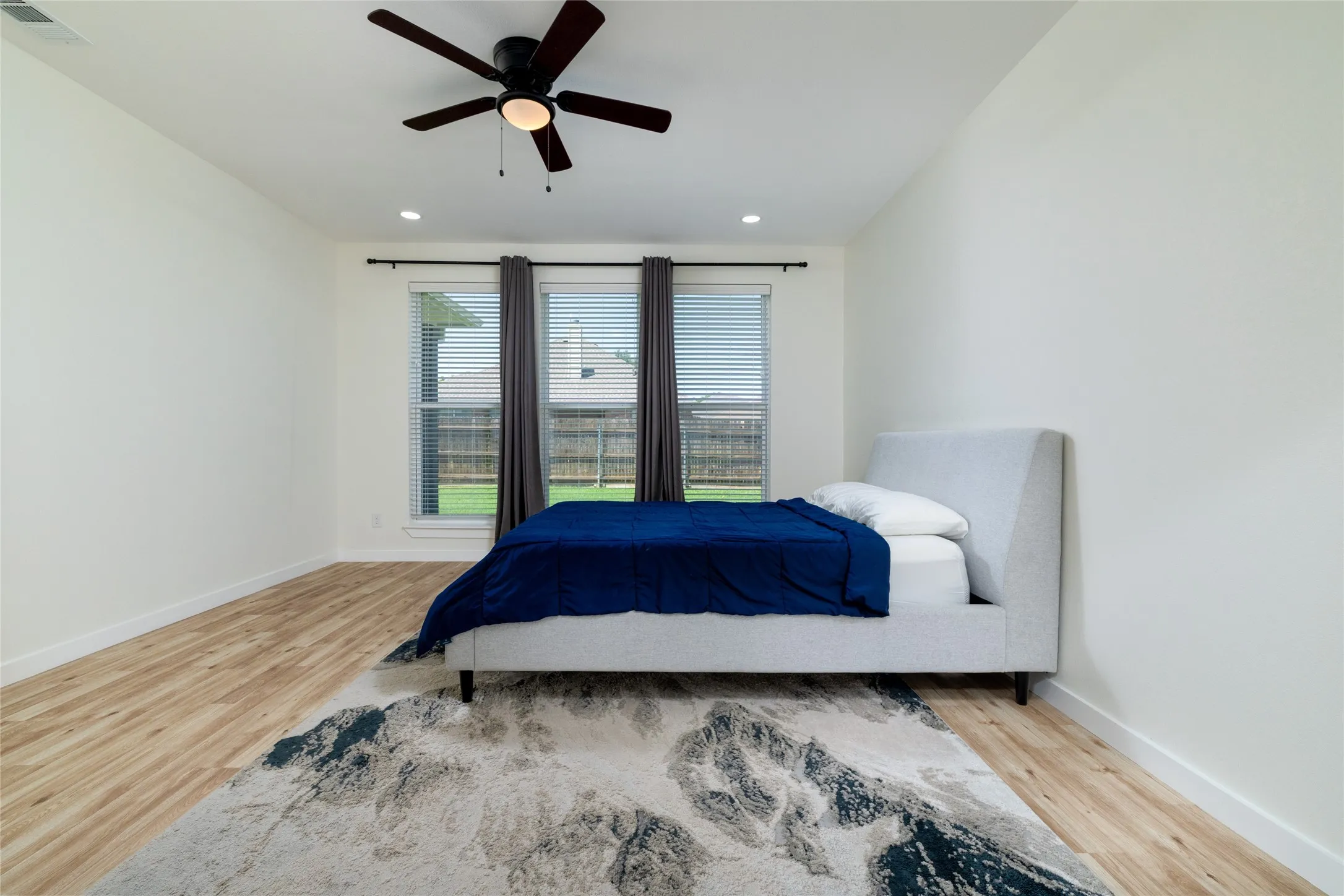 Original Photo of AI Photo - Spacious room featuring wood-finish flooring, a contemporary ceiling fan, recessed lighting, and large windows with blinds and drapes