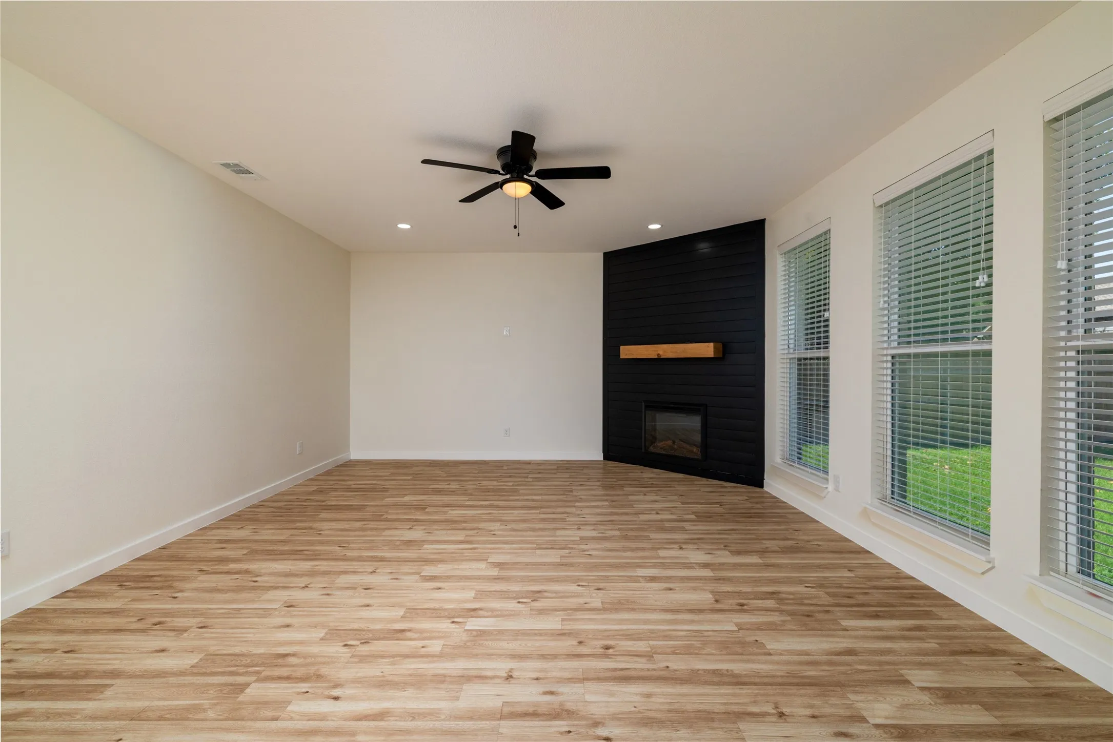 Original of AI photo - Spacious room featuring wood-finish flooring, a contemporary fireplace with a wood mantle, and multiple windows with blinds