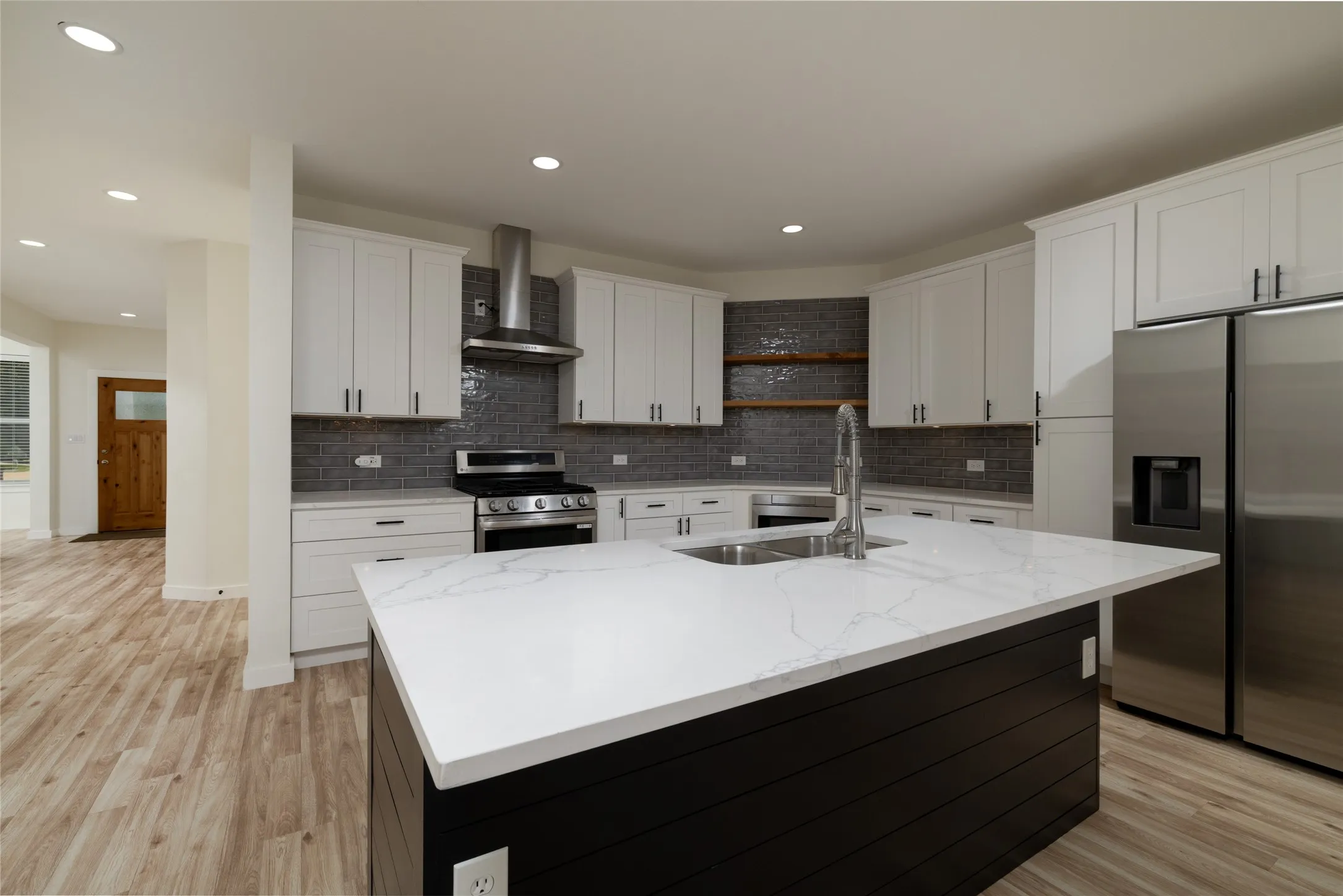 Modern kitchen featuring a large central island with a white countertop, white cabinetry with black hardware, stainless steel appliances, and wood-finish flooring