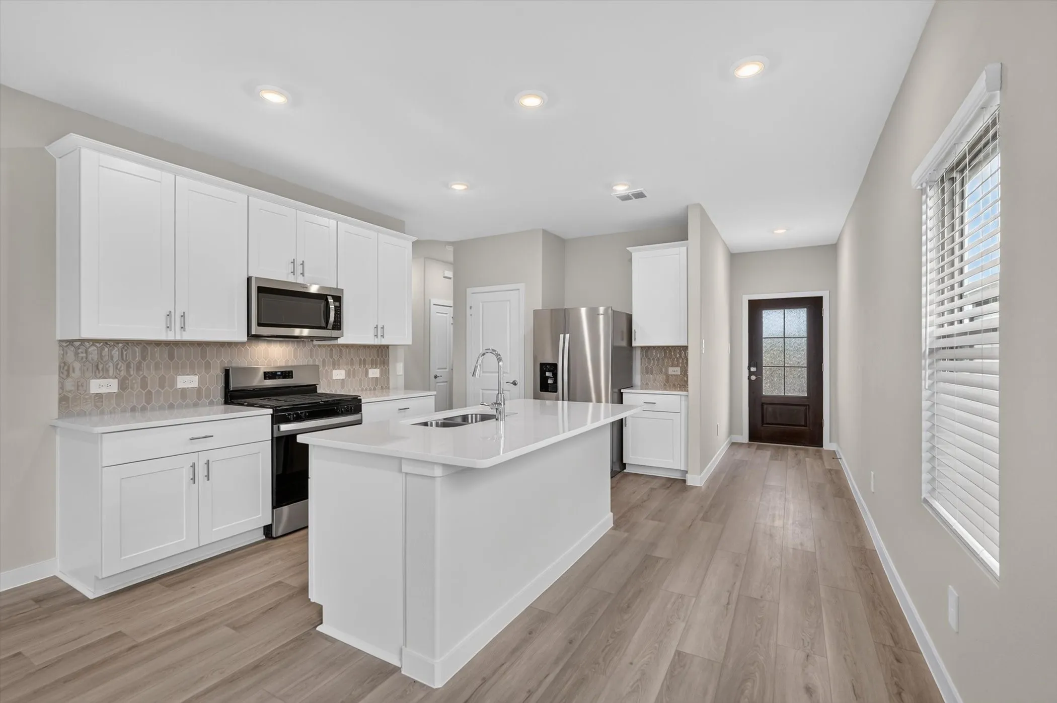Kitchen featuring white shaker cabinetry, stainless steel appliances, a center island with a sink, quartz countertops, and wood-finish flooring