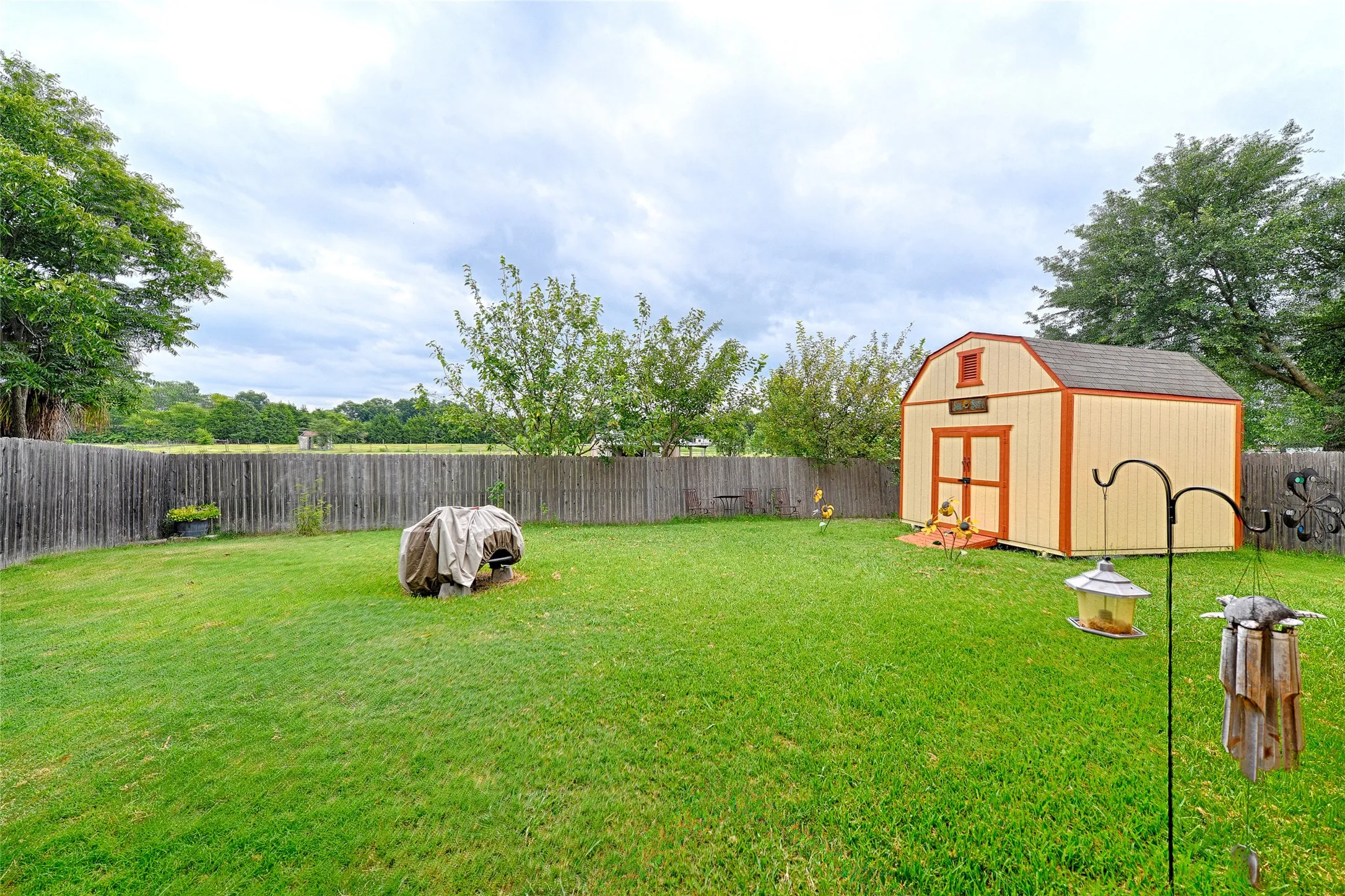 Expansive green lawn featuring a wooden privacy fence, mature trees, and a detached storage shed with double doors and a gambrel roof