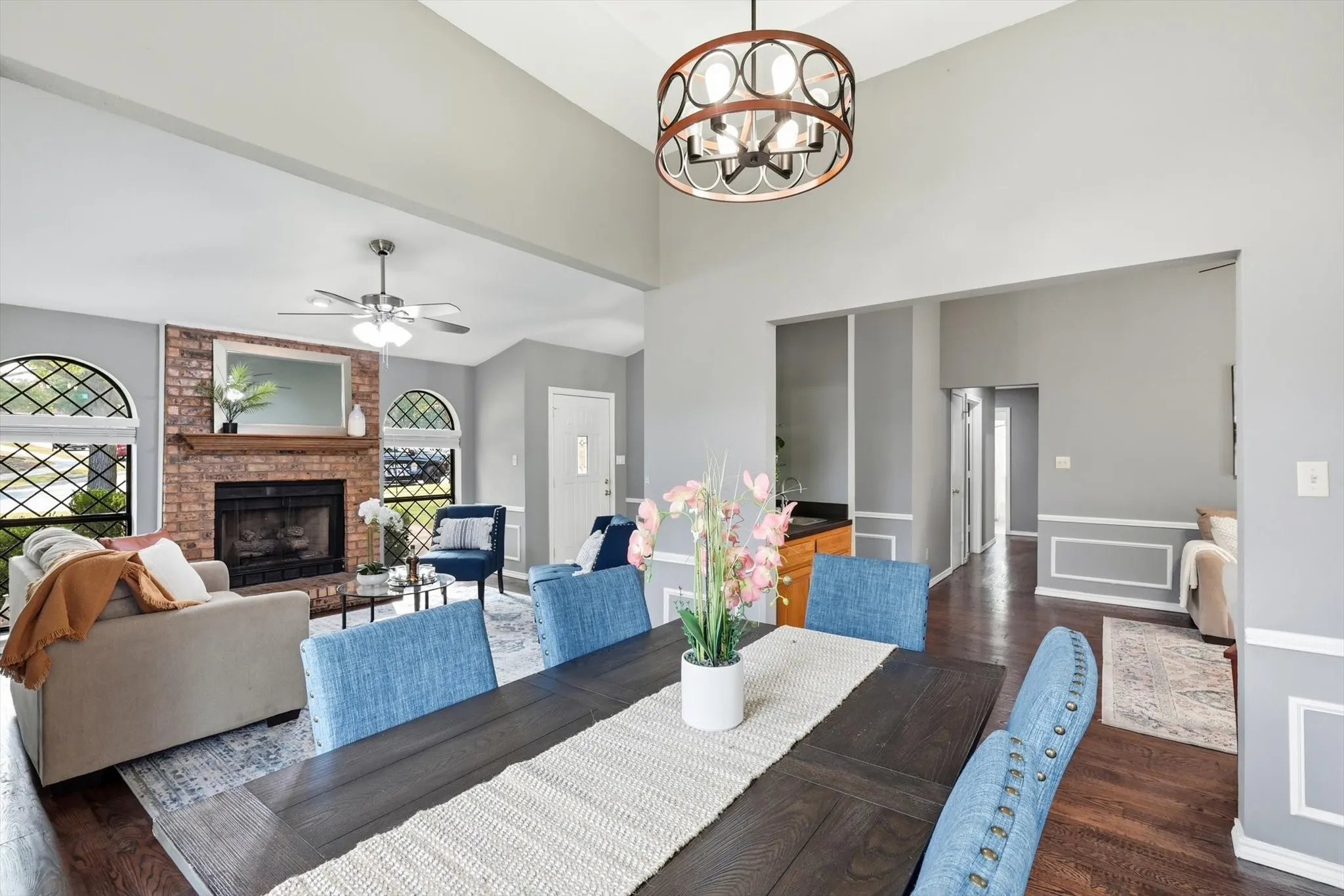 Open-concept living and dining area featuring dark wood-finish flooring, a linear dining table with seating for six, and expansive sight lines to a brick fireplace with decorative surround