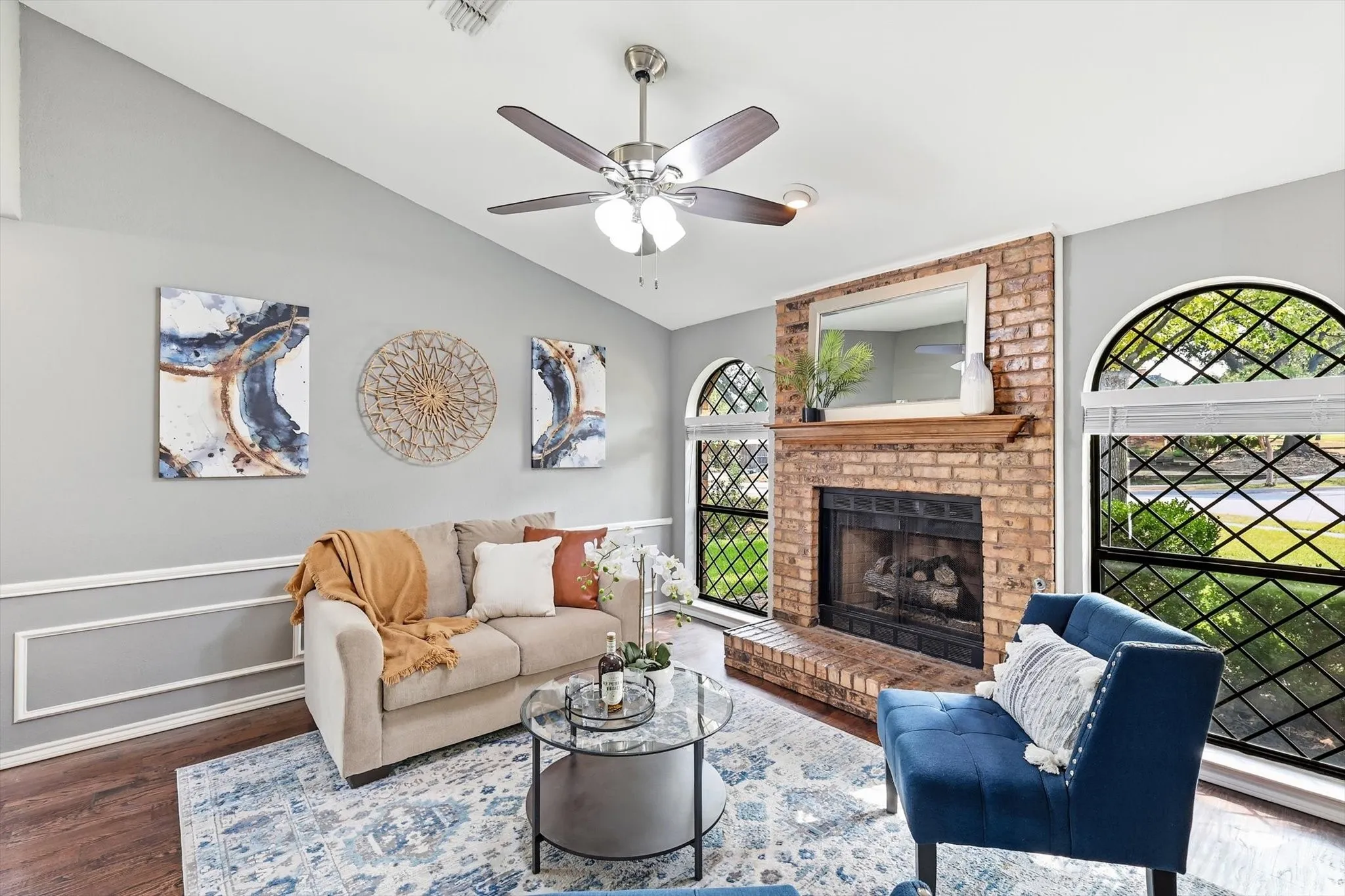 Elegant living room showcasing a brick fireplace with dark metal surround and glass doors, hardwood flooring, ceiling fan with integrated lighting, and decorative wall molding