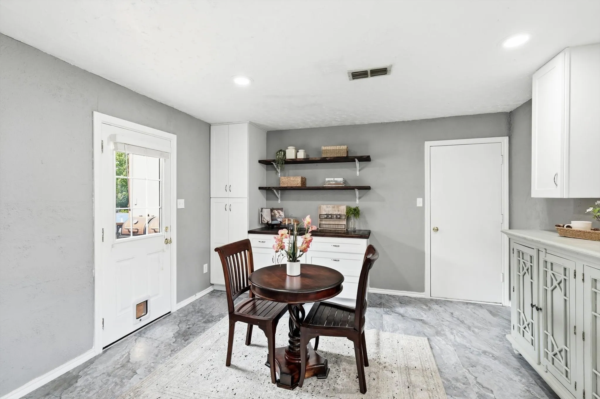 Elegant dining area showcasing soft gray walls, open floating shelving with decorative display space, recessed ceiling lighting, and light-toned wood-finish flooring
