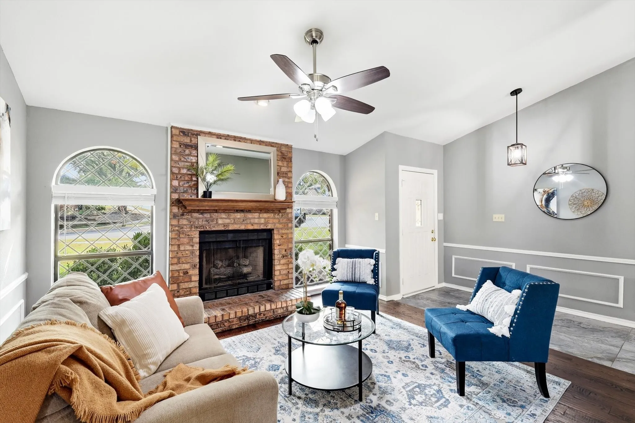 Bright living room anchored by a brick fireplace with dark surround and mantel, featuring arched windows framing natural landscape vistas, hardwood flooring, ceiling fan with integrated lighting, and recessed wall paneling detail