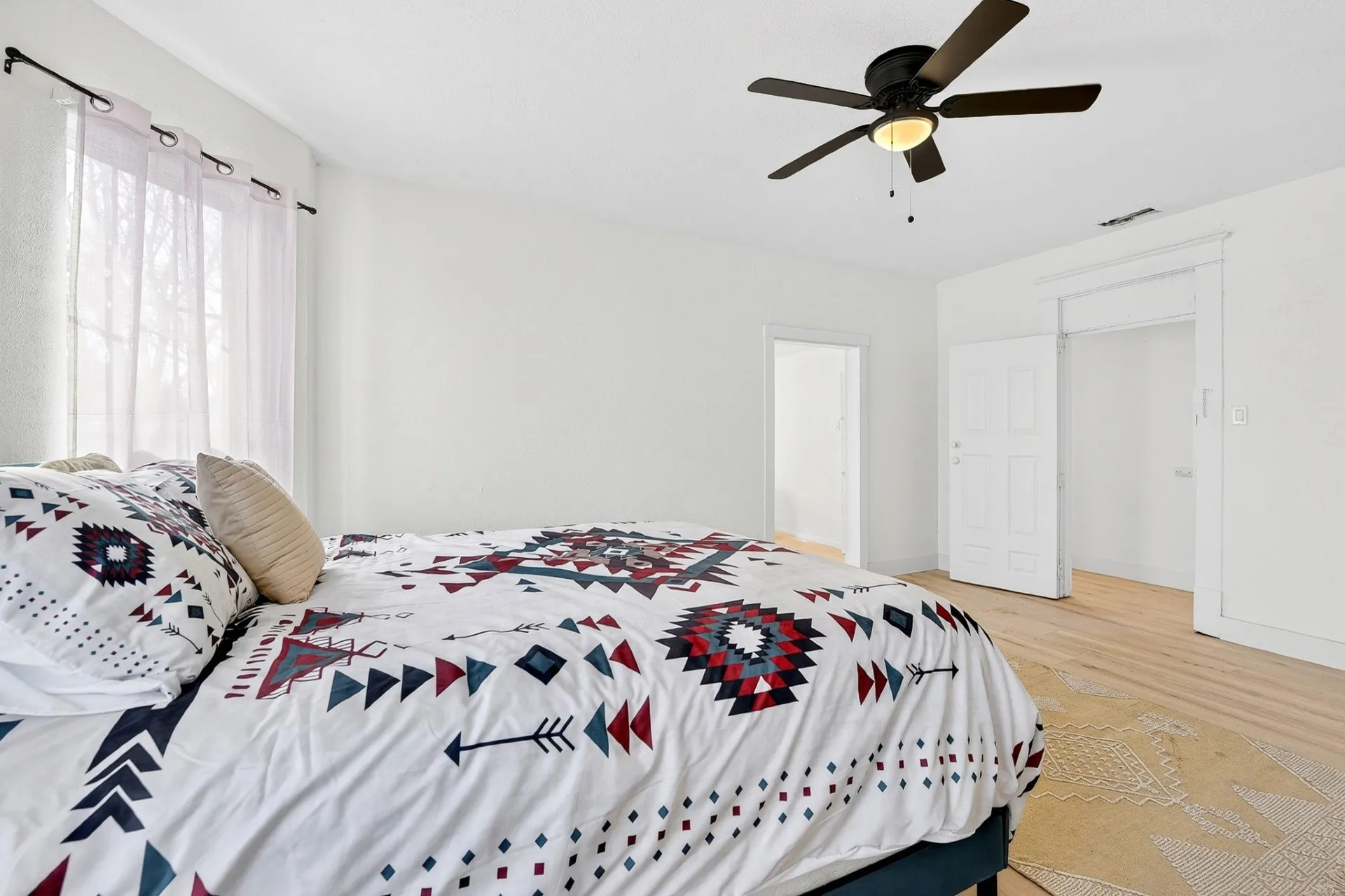 Bedroom with light wood-finish flooring, a ceiling fan with integrated lighting, and a window featuring sheer drapery