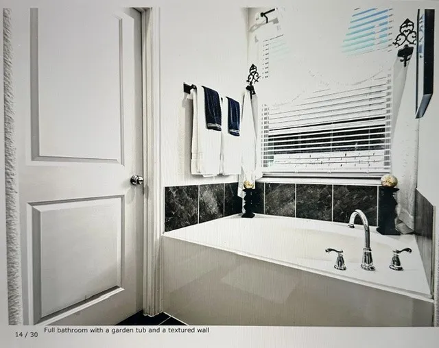 Garden tub with deck-mounted chrome fixtures, surrounded by dark tile accents and a window with horizontal blinds