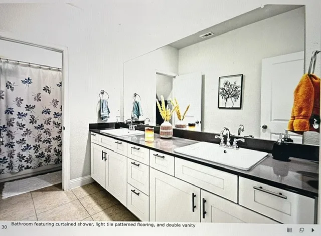 Bathroom featuring a double vanity, dark countertop, dual chrome faucets, white cabinetry with dark hardware, and light-toned tile flooring