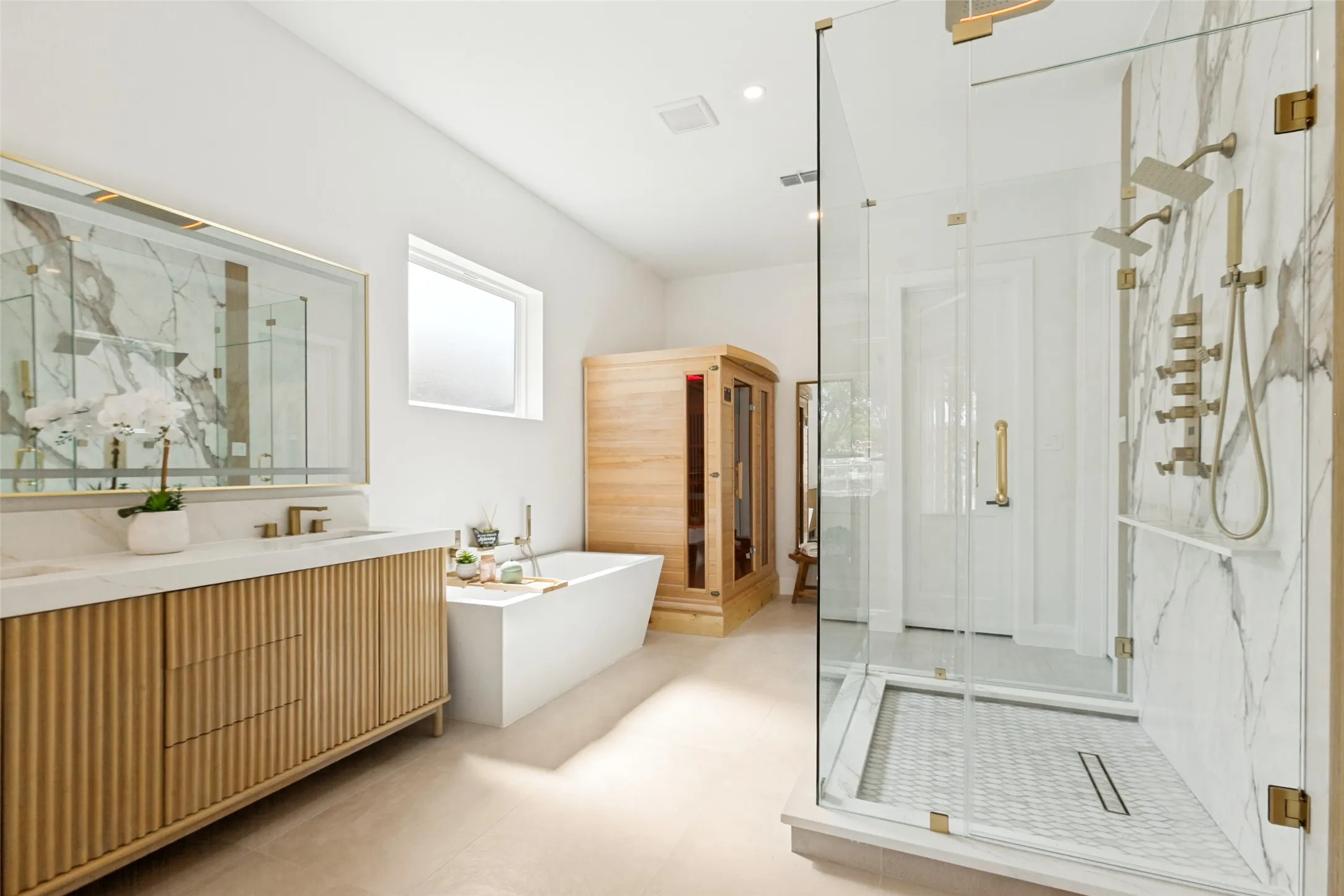Primary Spa Bathroom with jetted freestanding soaking tub, dual fluted wood vanity, dual walk-in shower with multiple shower heads,overhead rain shower and infrared therapy sauna. Designer quartz and marble-patterned tile in shower, and glass shower enclosure.