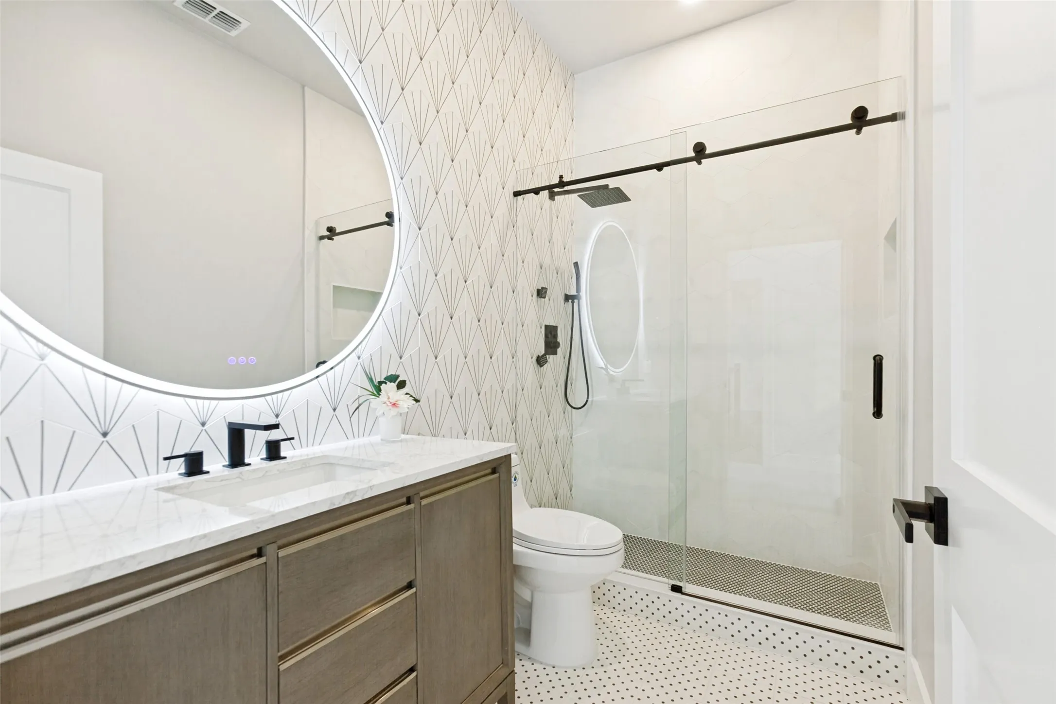 Lighted circular vanity mirror, wood vanity with quartz top, sleek matte black fixtures, glass-enclosed shower with rain shower head, and Designer tile walls and flooring.