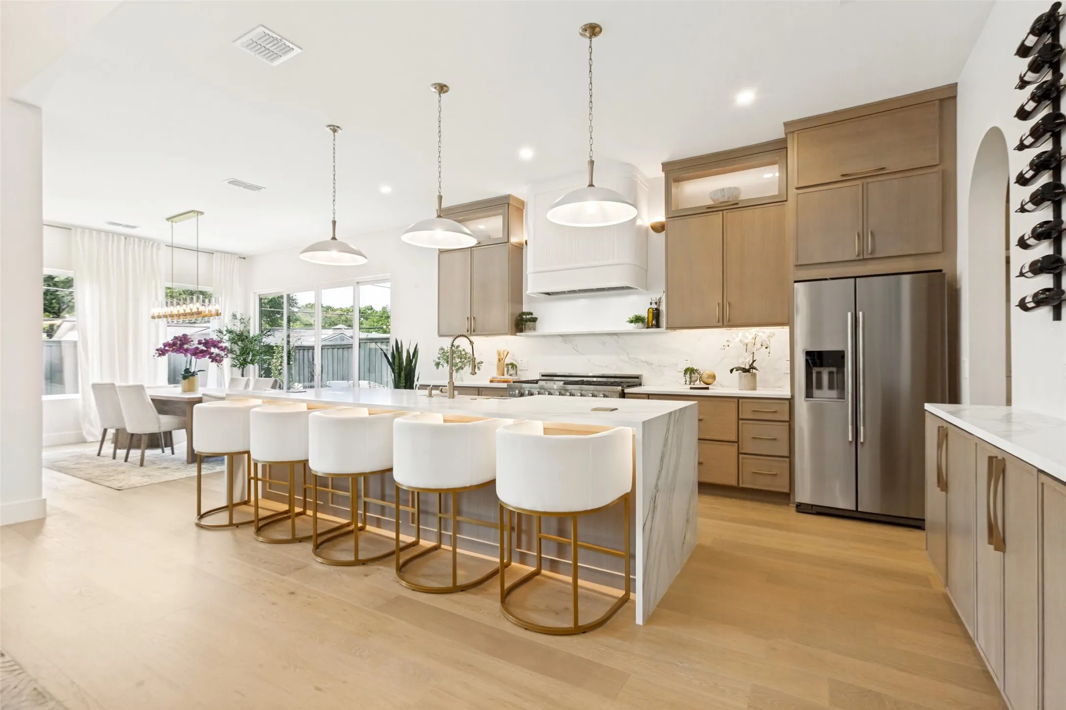 Modern chef's kitchen featuring white oak wood soft close cabinetry, stainless steel appliances, quartz waterfall-edge island with seating for multiple guests, designer pendant lighting, and expansive oak wood flooring throughout the open-concept living area with abundant natural light from large windows.