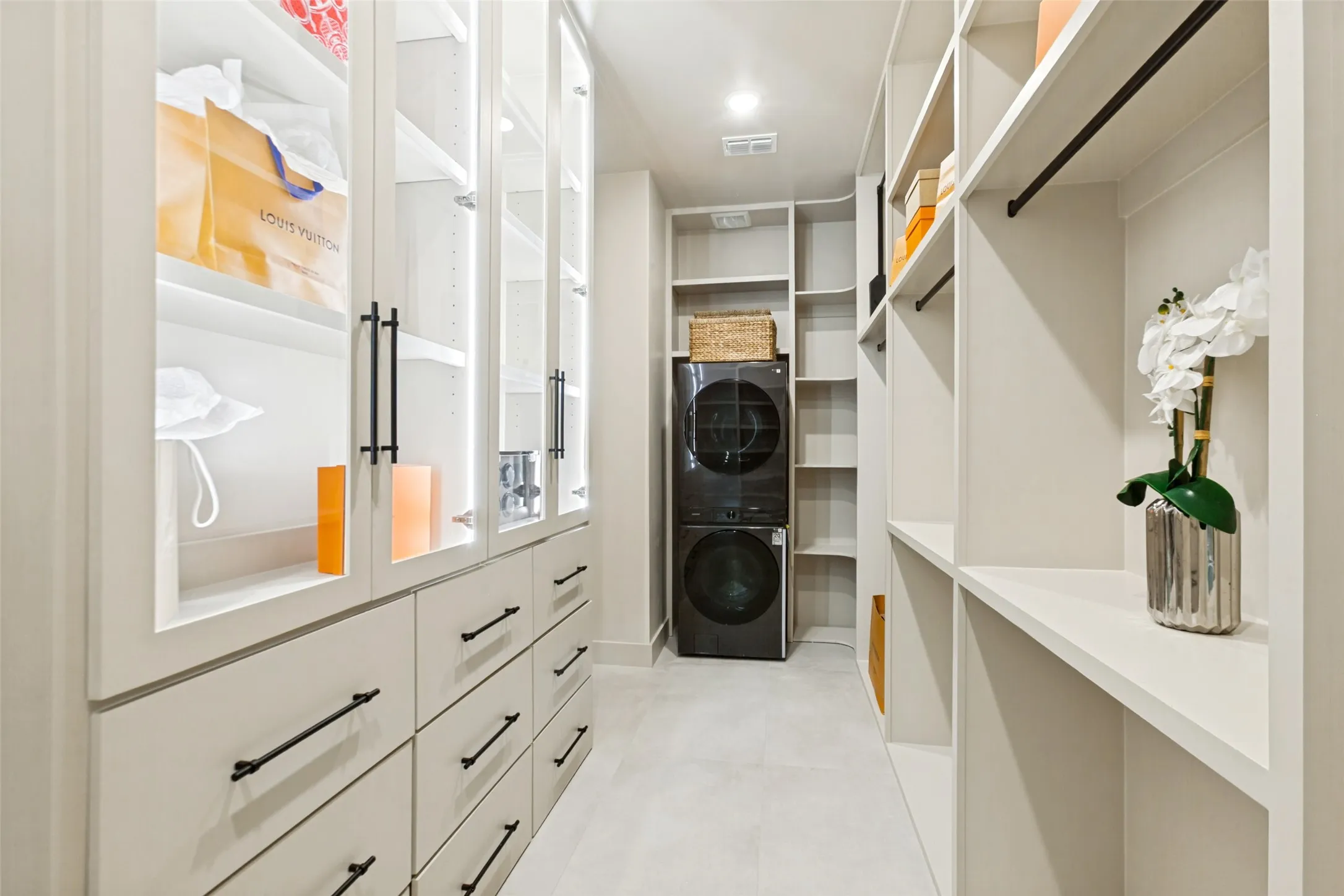 Custom closet system featuring integrated lighting, glass-front cabinetry, drawers with designer gun-metal hardware, designer pull down wardrobe and extensive open shelving. Stacked washer and dryer.