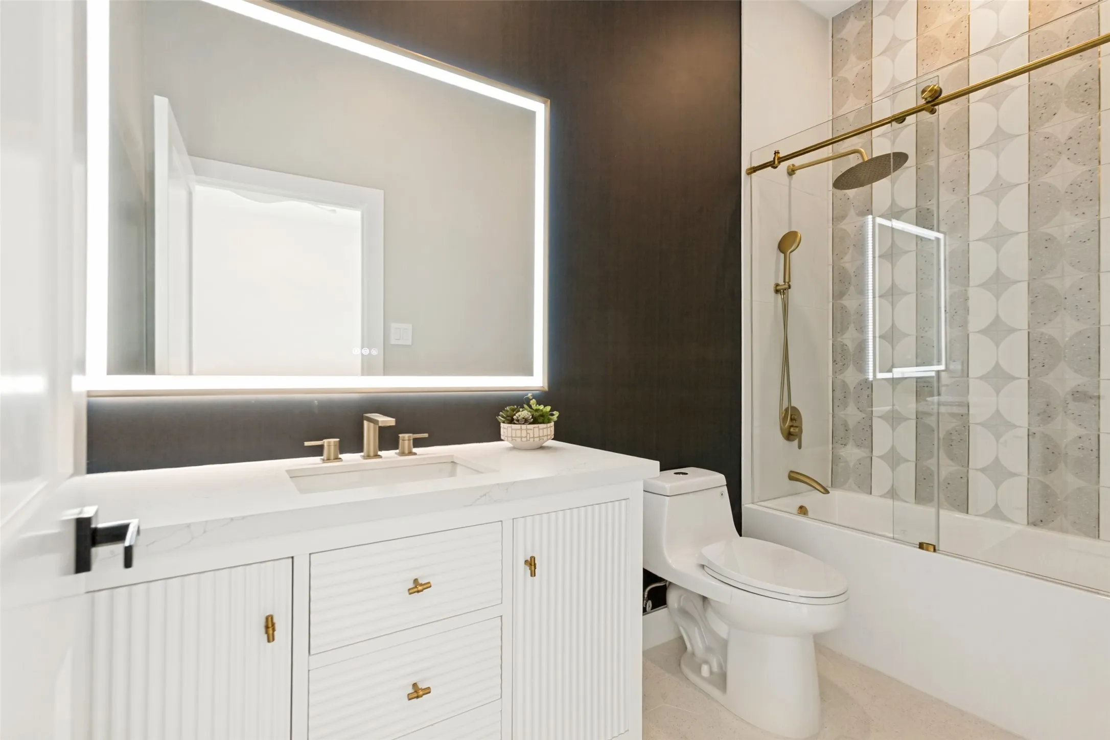 Bathroom to bedroom #5 with backlit vanity mirror, fluted vanity with quartz top, gold fixtures, designer tile, and a glass shower enclosure with gold accents.