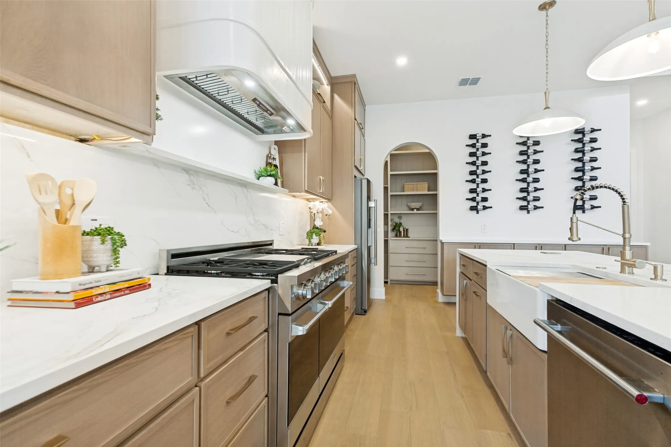 Modern kitchen featuring white oak wood cabinetry, quartz countertops, stainless steel gas range with sculptural hood, and a farmhouse-style apron sink with contemporary faucet.