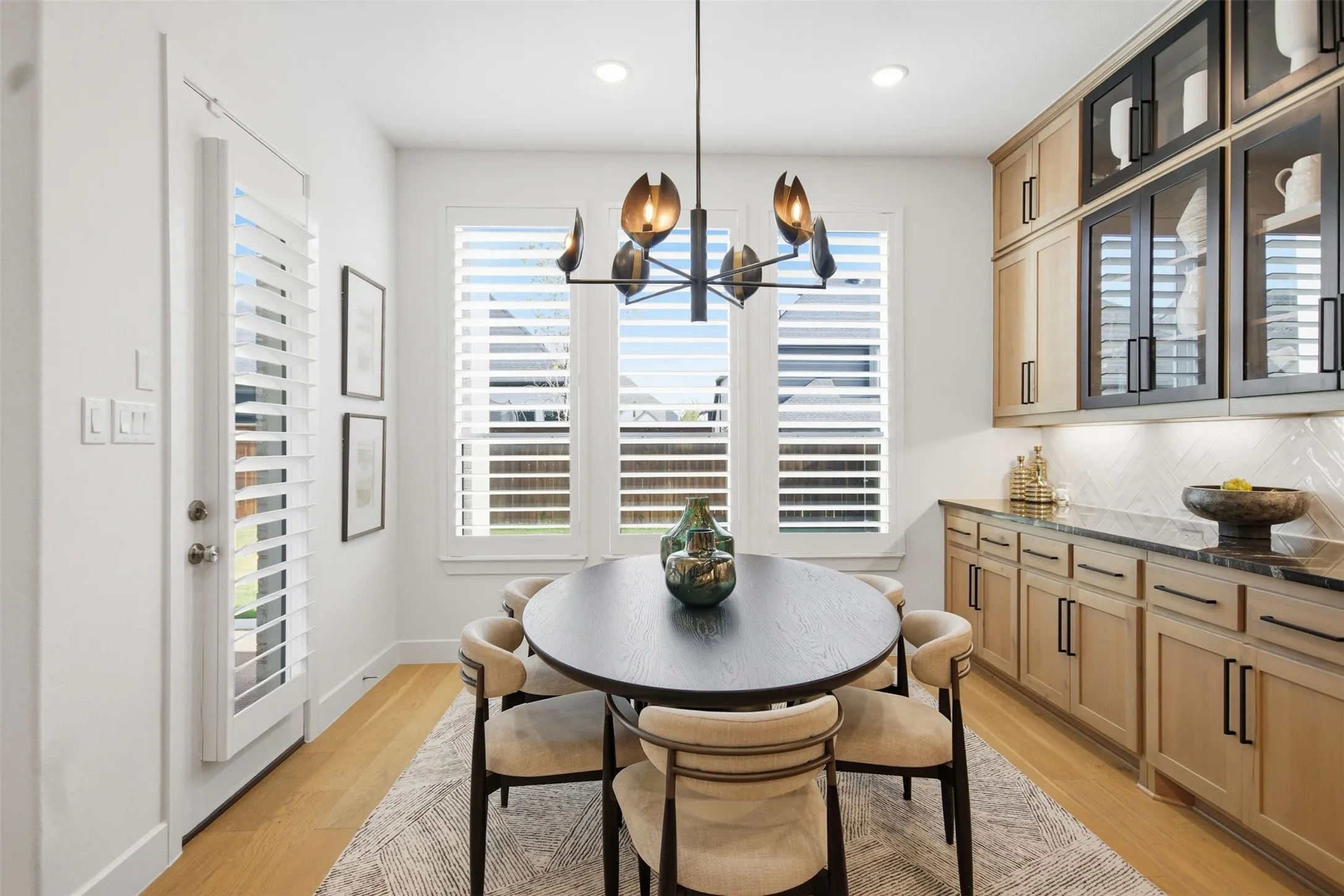 Dining area featuring three large windows with plantation shutters, a contemporary chandelier, a built-in buffet with dark countertops, wood-finish flooring, and glass-front upper cabinetry