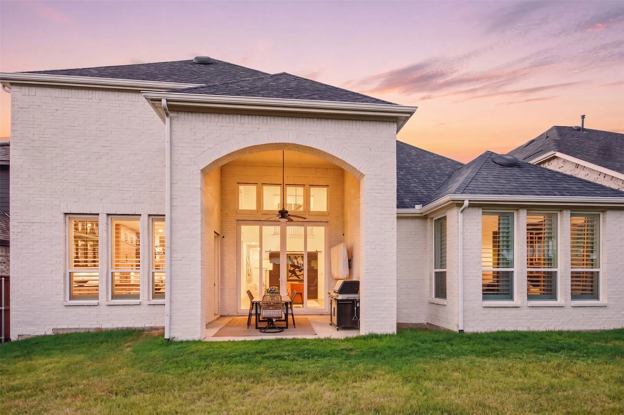 Expansive rear exterior featuring white brick siding, a covered patio with an arched entry, a ceiling fan, and large windows with plantation shutters