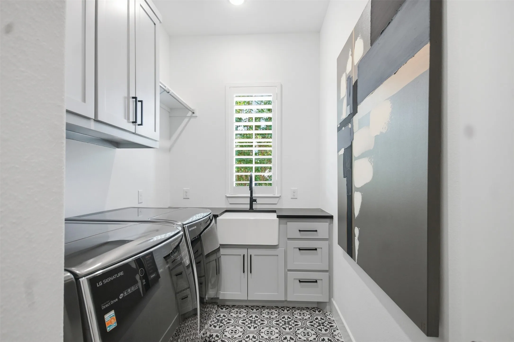 Utility space featuring patterned floor tile, light cabinetry with dark hardware, a single basin sink with a dark faucet, dark countertops, and a window with plantation shutters
