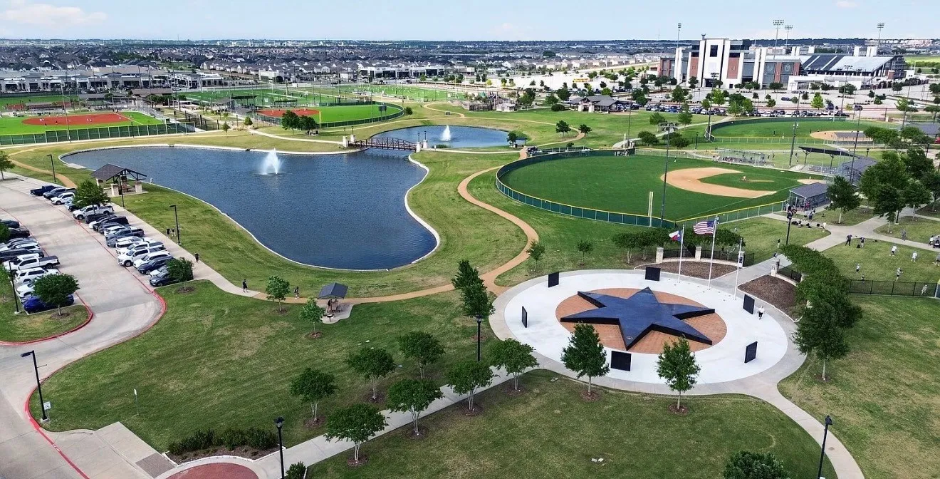 Expansive park featuring a large pond with fountains, multiple sports fields including a baseball diamond, walking paths, and a prominent star-shaped monument