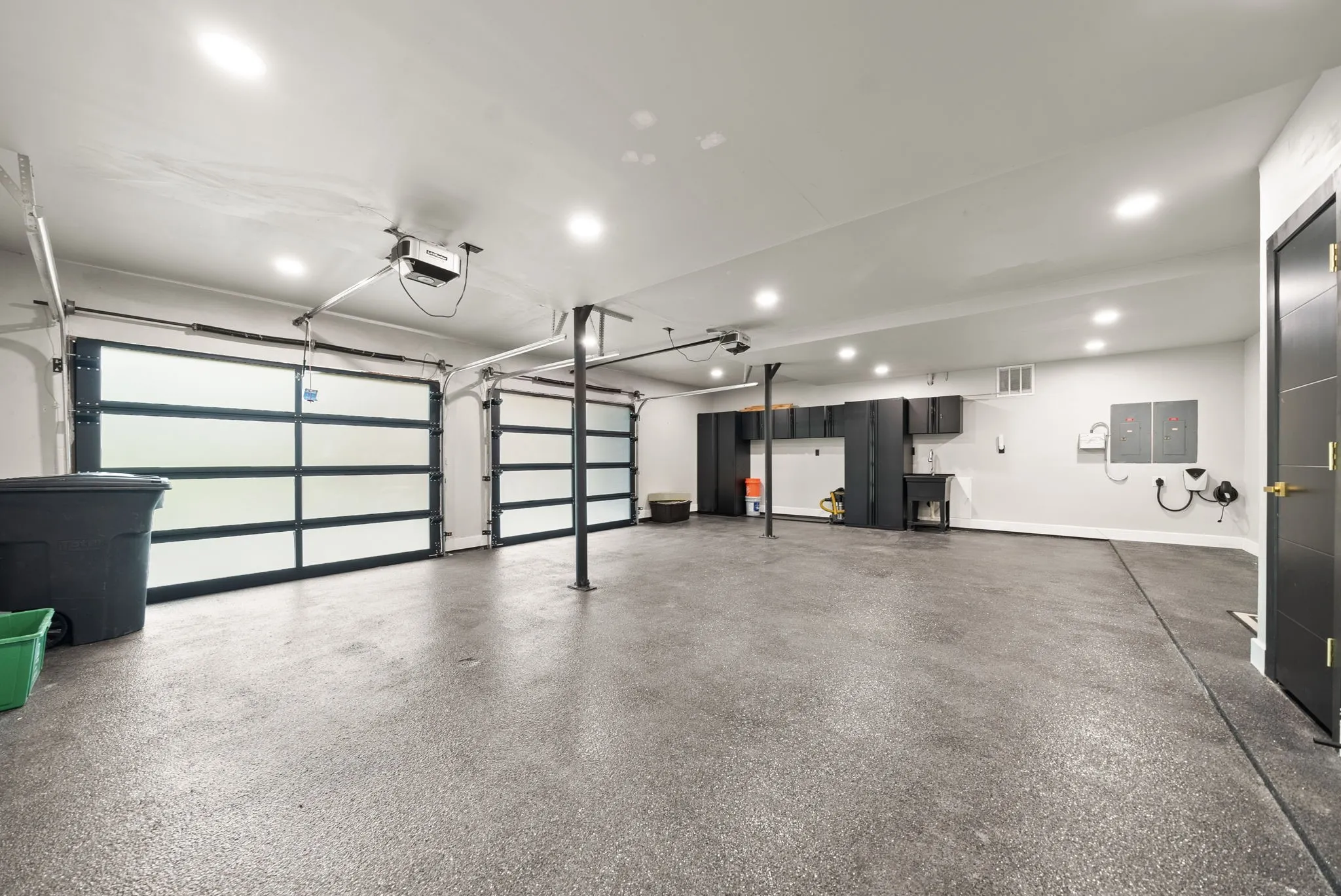 Expansive garage featuring two modern glass-paneled garage doors, a speckled epoxy floor coating, recessed lighting, built-in dark cabinetry with integrated sink, and an electric vehicle charging station