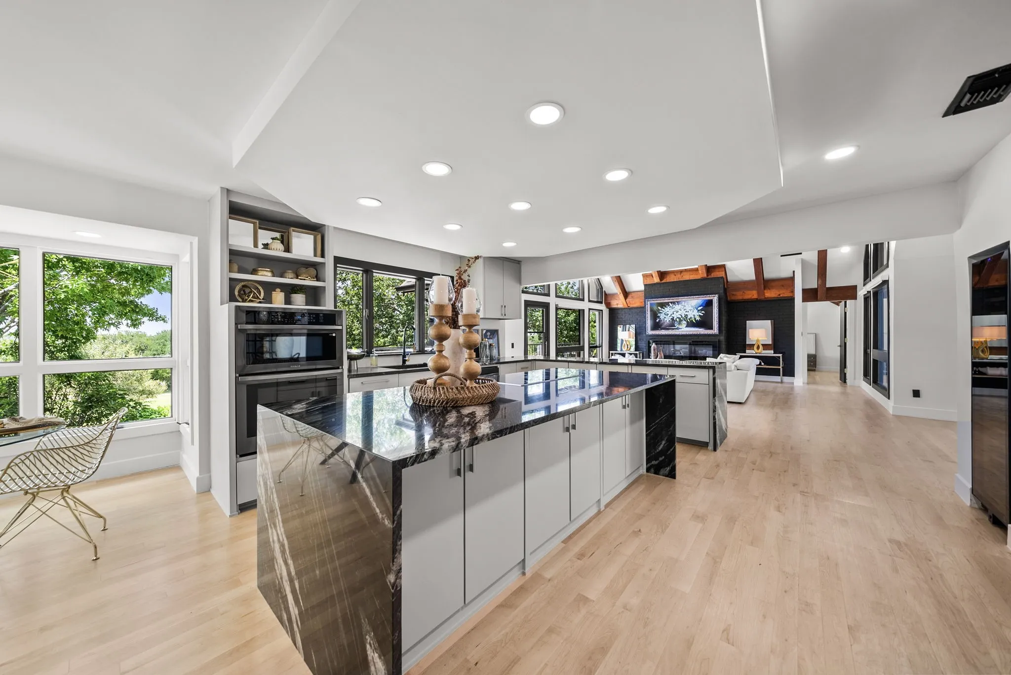 Spacious open-concept kitchen featuring a large island with a waterfall-edge countertop, light wood-finish flooring, recessed lighting, built-in double ovens, and ample natural light from multiple windows