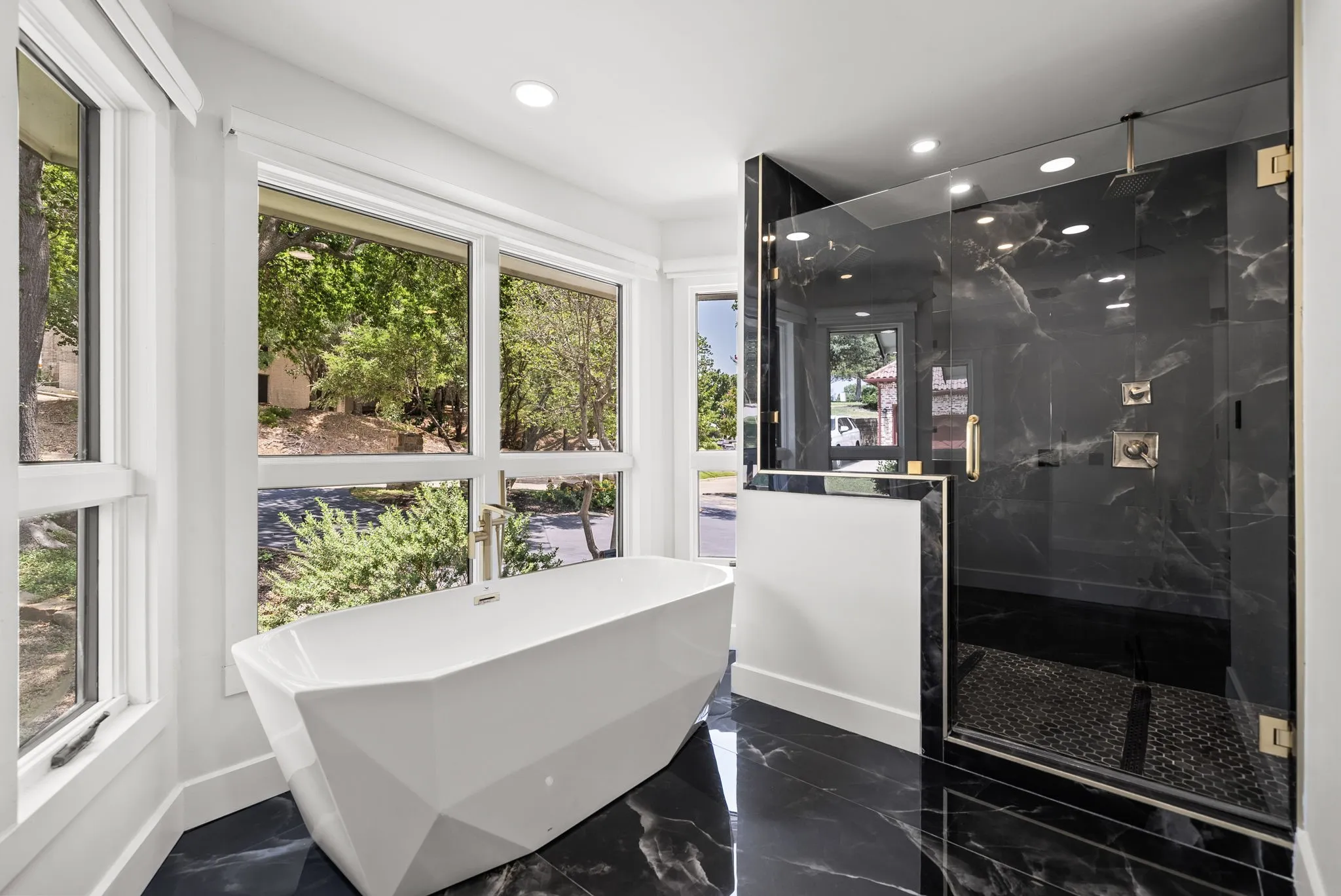 Contemporary bathroom featuring a geometric freestanding tub, expansive windows, a spacious glass-enclosed shower with black marble-style tiling, gold-tone fixtures, and glossy black flooring