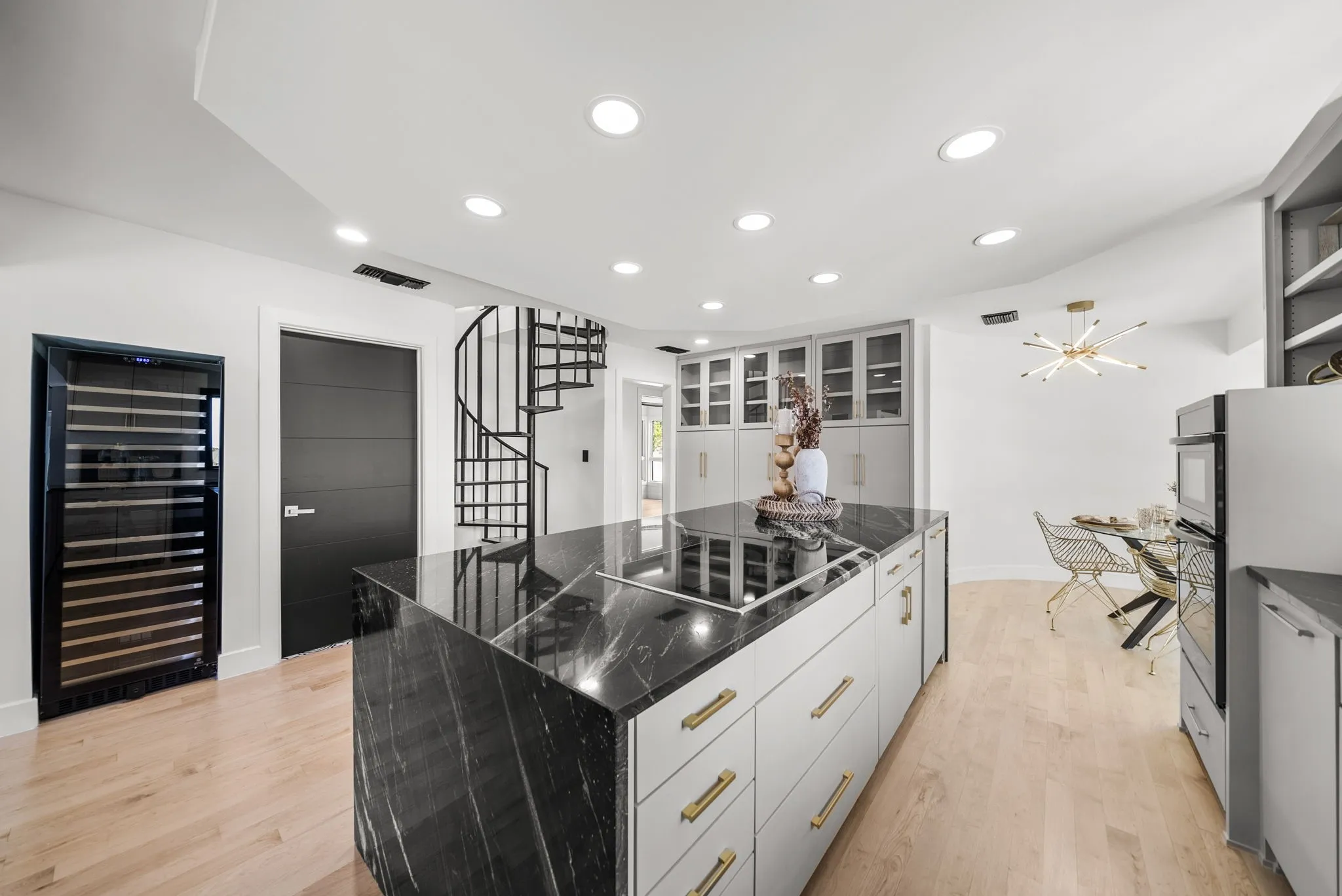 Contemporary kitchen island featuring a black marble countertop with integrated cooktop, light wood-finish flooring, recessed lighting, a built-in wine cooler, and a spiral staircase