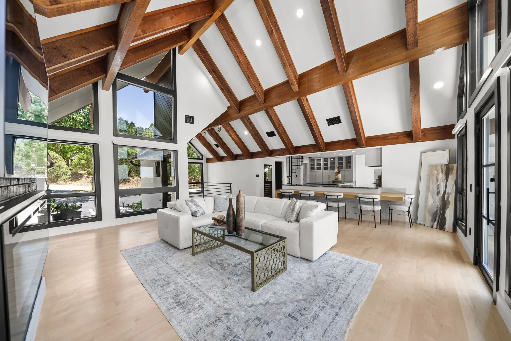 Vaulted ceiling with exposed wood beams, expansive window array, light wood-finish flooring, recessed lighting, and a kitchen featuring a peninsula with bar seating