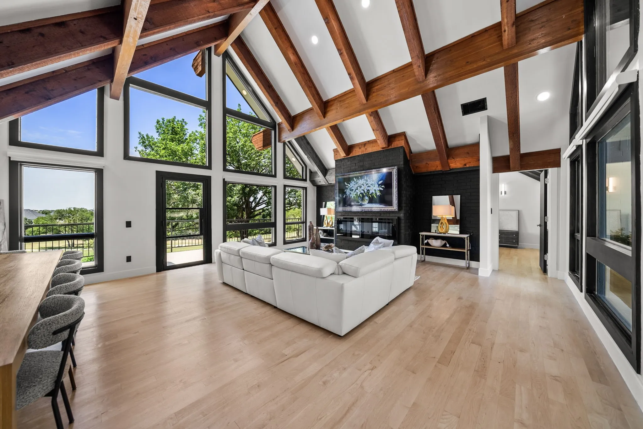 Vaulted great room featuring exposed wood beams, expansive floor-to-ceiling windows, light wood-finish flooring, a contemporary fireplace with a large integrated television, and a built-in bar with seating