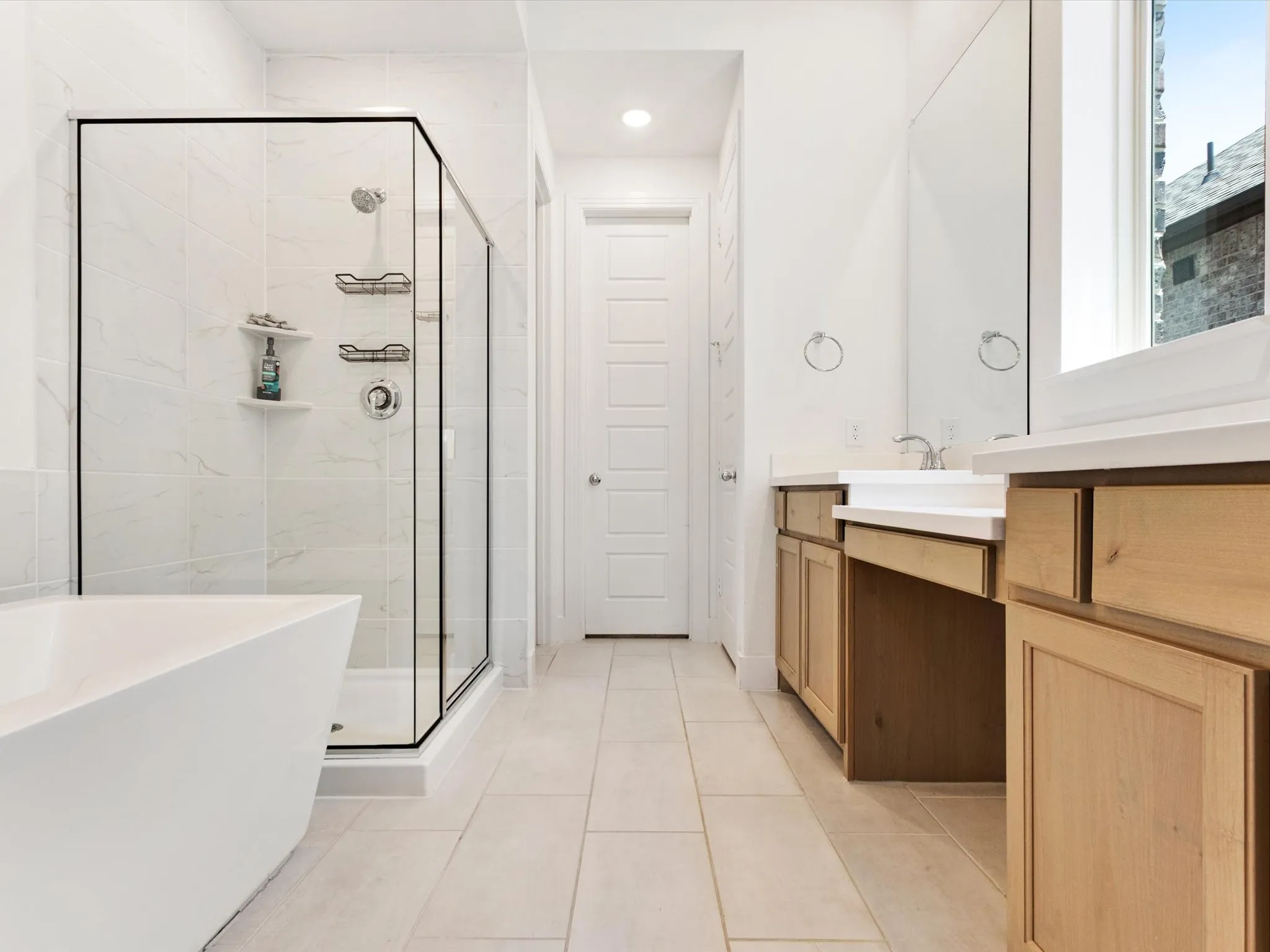 Contemporary bathroom featuring a freestanding soaking tub, a frameless glass shower enclosure with white tile surround, a double vanity with light wood cabinetry and white countertops, and a large mirror