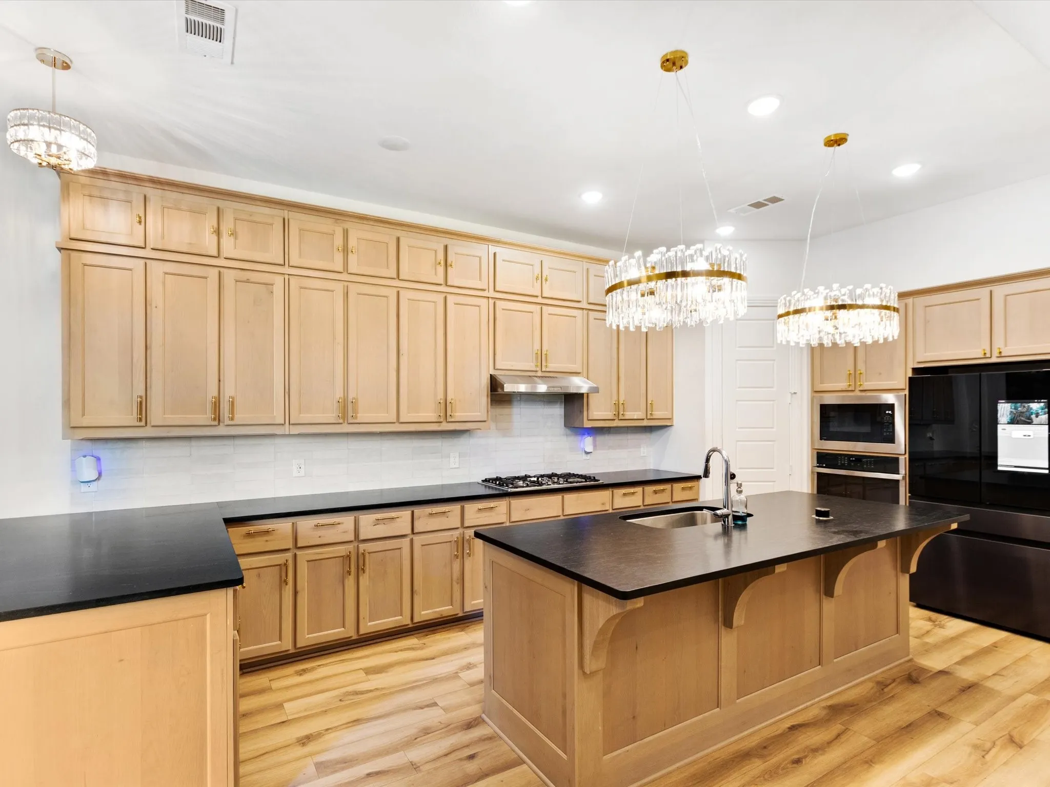 Kitchen featuring light wood cabinetry with gold-toned hardware, dark countertops, a white subway tile backsplash, and wood-finish flooring