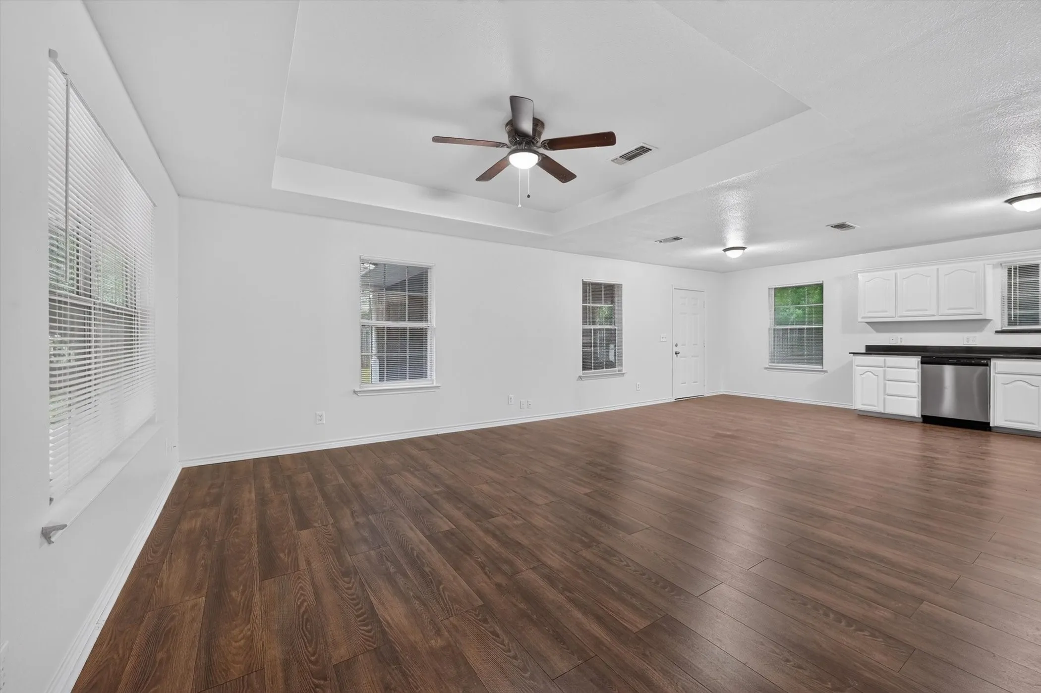 Expansive open-concept room featuring wood-finish flooring, a tray ceiling with a ceiling fan, multiple windows with blinds, recessed lighting, and a kitchen area with white cabinetry and a stainless steel dishwasher